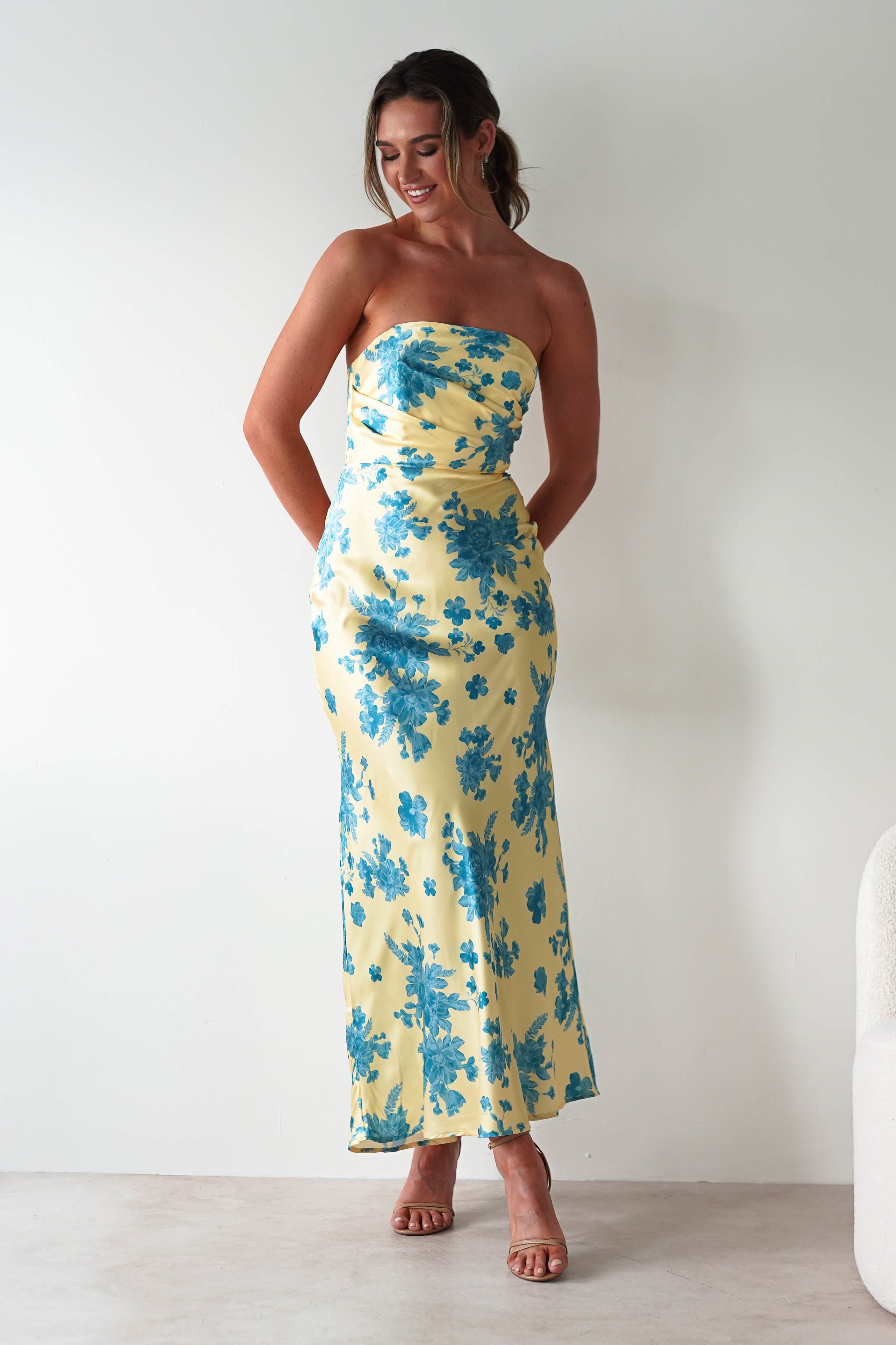 A woman stands against a light wall, wearing a strapless satin maxi dress in yellow with blue floral patterns and nude heels. She smiles and looks down, hands behind her back—a perfect wedding guest dress.