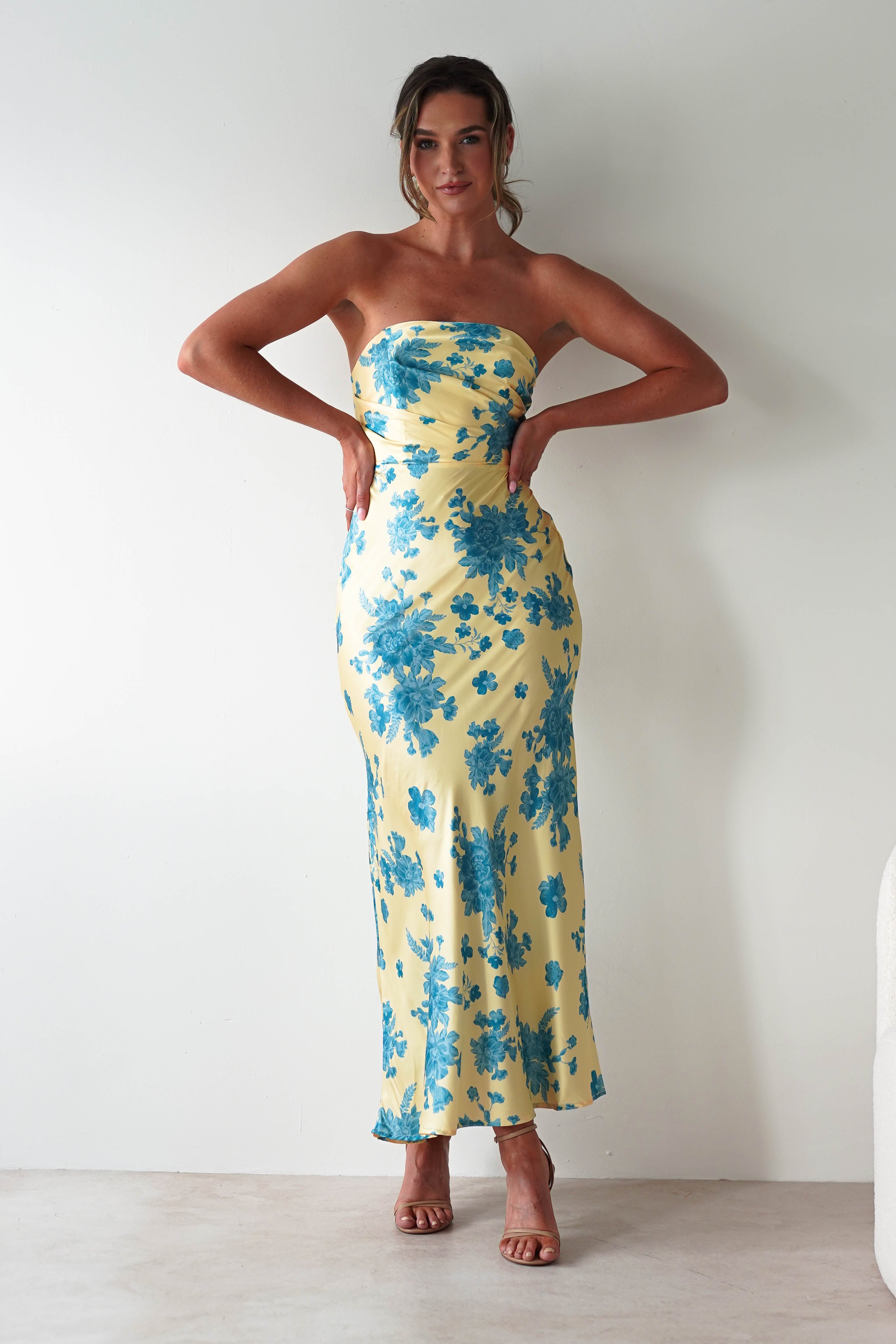 A woman stands against a plain white wall wearing a strapless satin maxi dress in yellow with blue floral patterns. She poses with her hands on her hips and finishes the look with brown high-heeled sandals, perfect as a stylish wedding guest dress.
