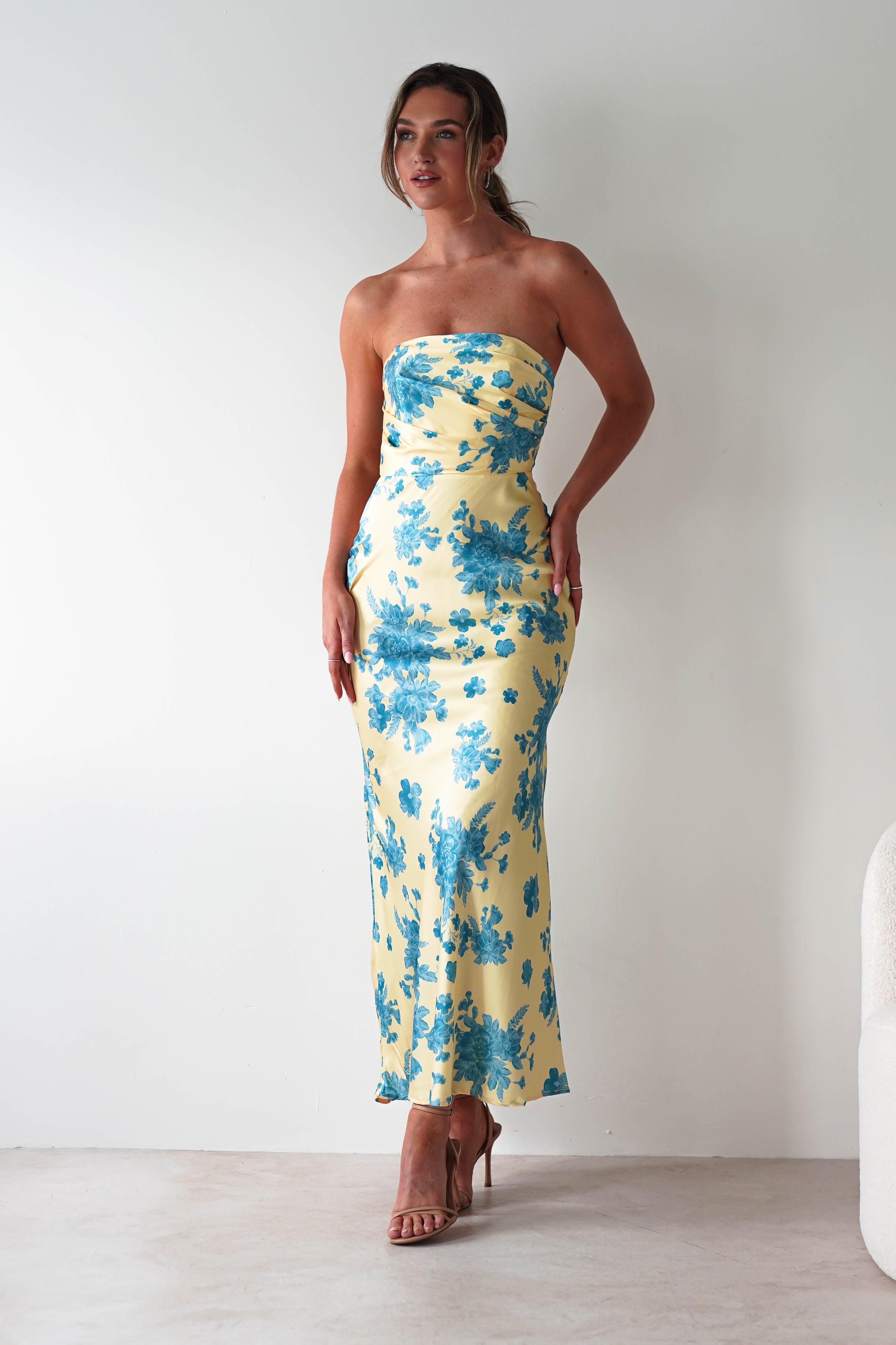 A woman stands against a plain white wall in a strapless satin maxi dress with yellow and blue floral patterns, paired with nude high-heeled sandals—a chic look perfect as a wedding guest dress.