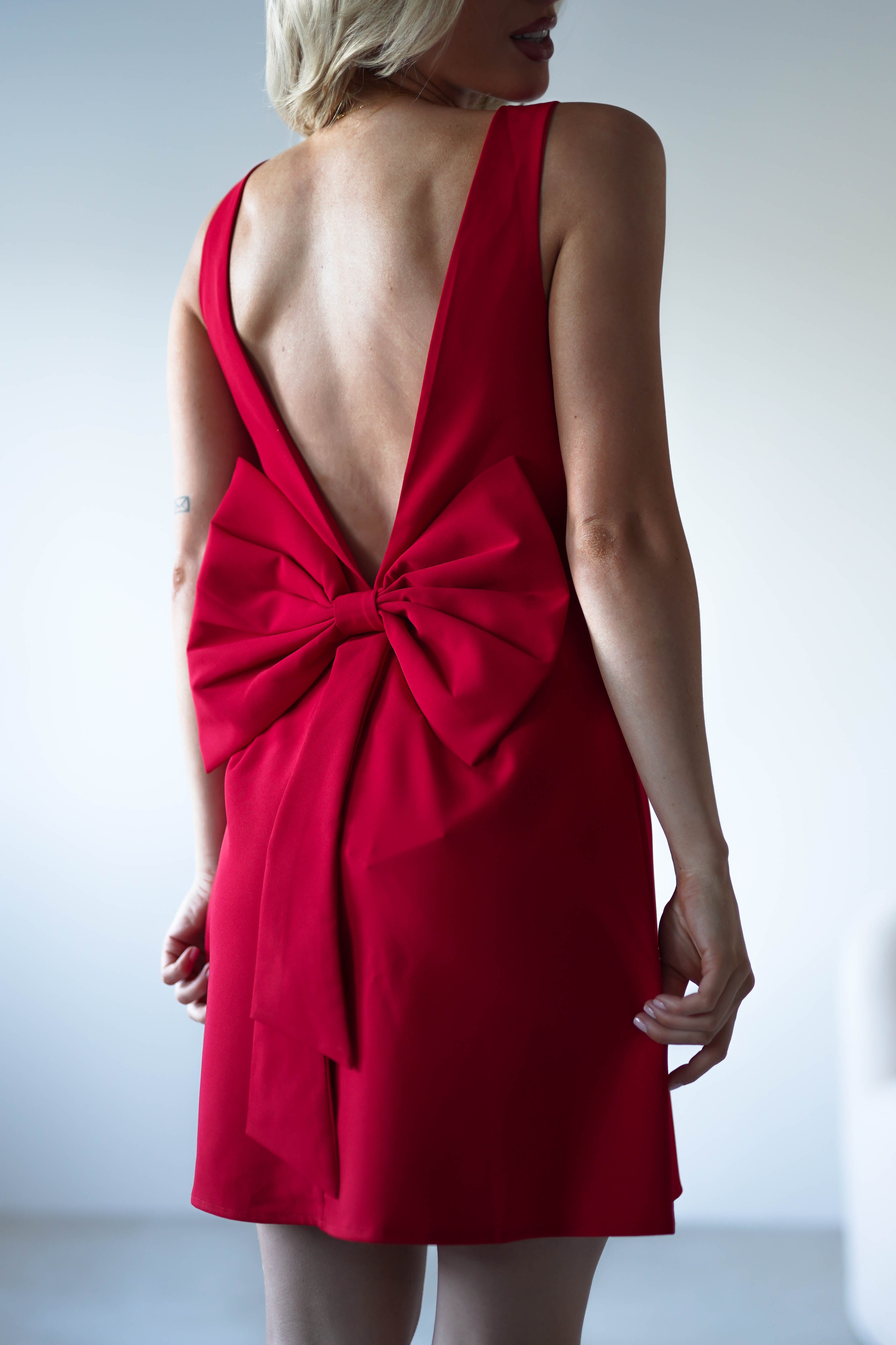 A woman wearing a sleeveless red mini dress with a deep V-back and a large decorative bow at the lower back stands against a plain light background—an elegant choice for an open back dress or wedding guest dress.