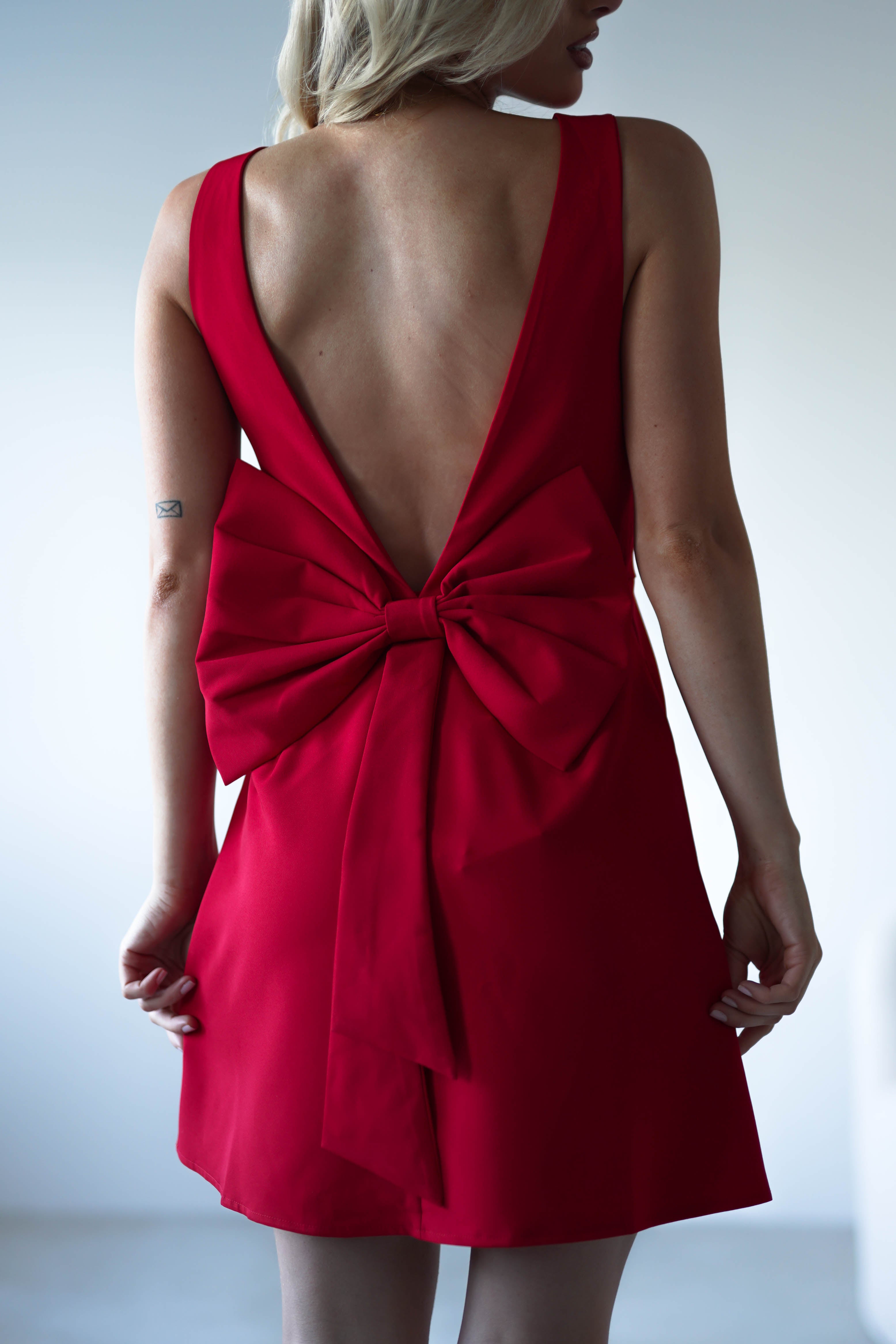 A woman wears a sleeveless red mini dress with a deep V-back, large bow detail at the lower back, and stands against a plain background—perfect as an elegant wedding guest dress.