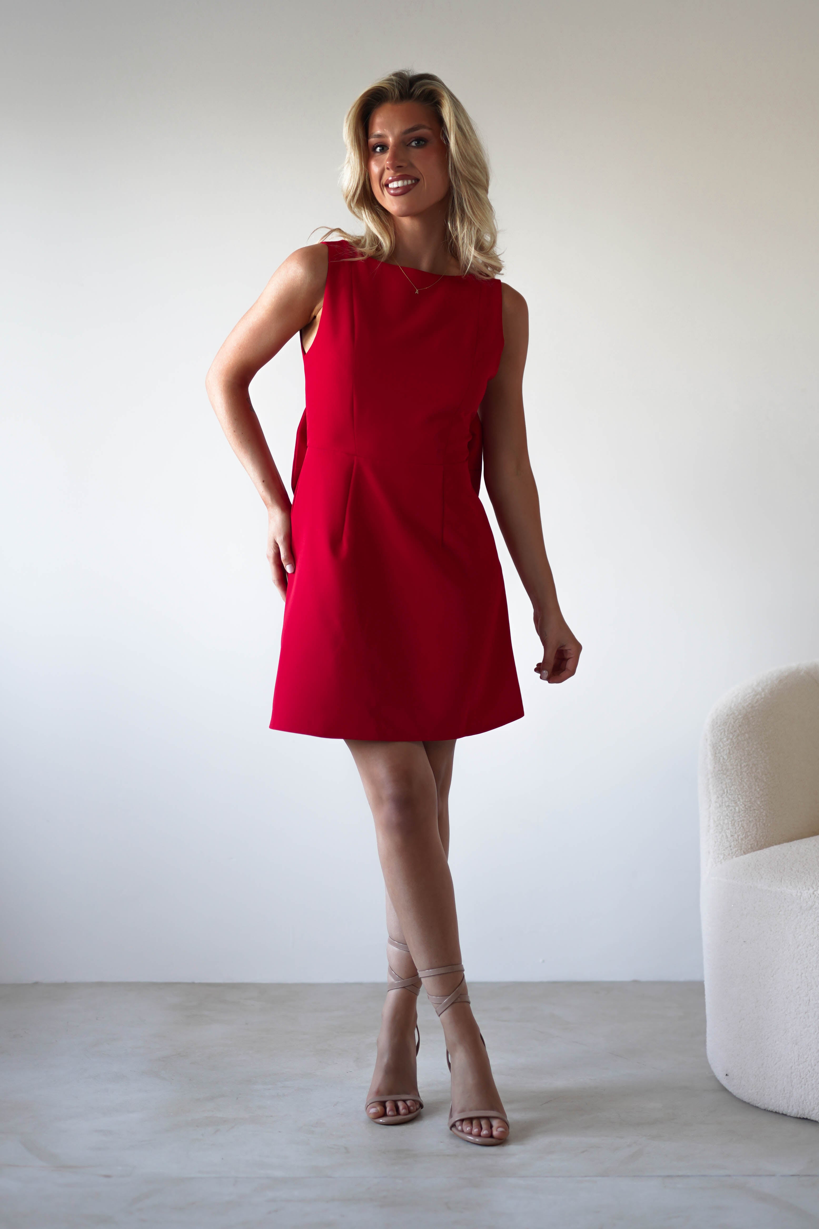 A woman with blonde hair wearing a sleeveless red mini dress and strappy nude heels stands indoors, smiling, against a plain light-colored background next to a textured chair—an ideal look for a wedding guest dress.
