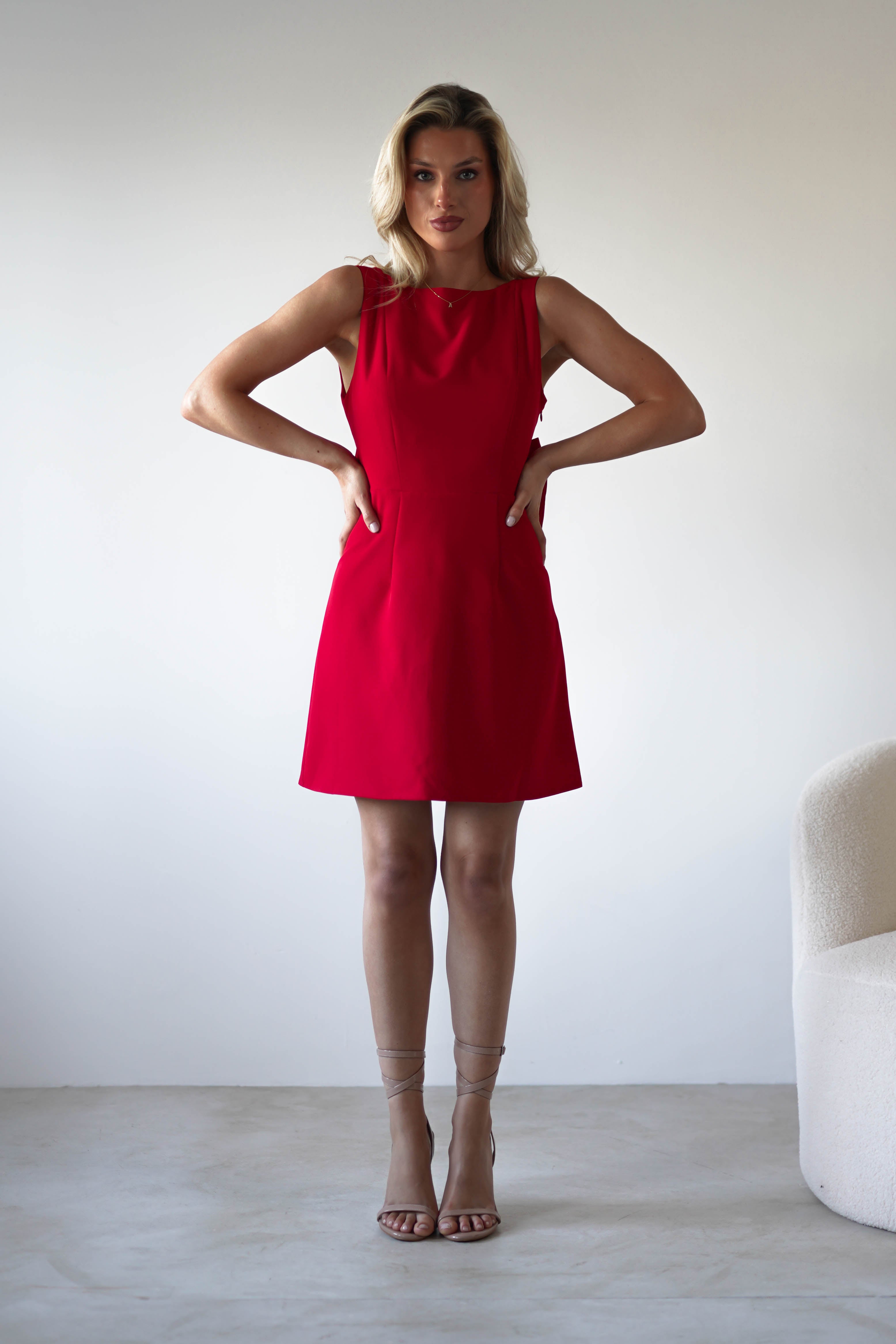 A woman stands indoors against a plain white wall, wearing a sleeveless red mini dress and strappy nude heels, with her hands resting on her hips. A white textured armchair is visible to the right.