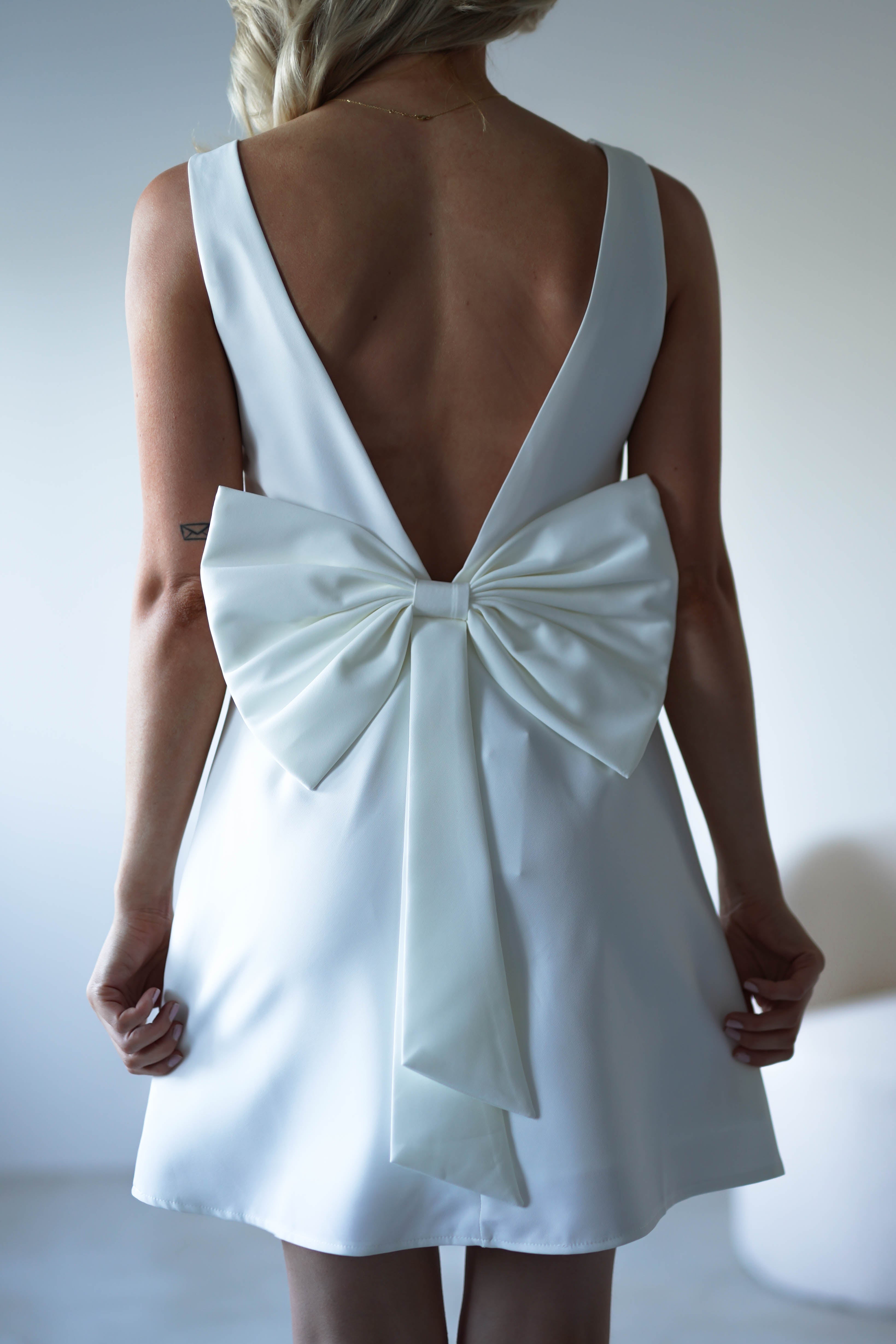 A woman wearing a sleeveless white mini dress with a deep V-back and a large white bow at the lower back, standing indoors and facing away from the camera—perfect as an elegant open back party dress.