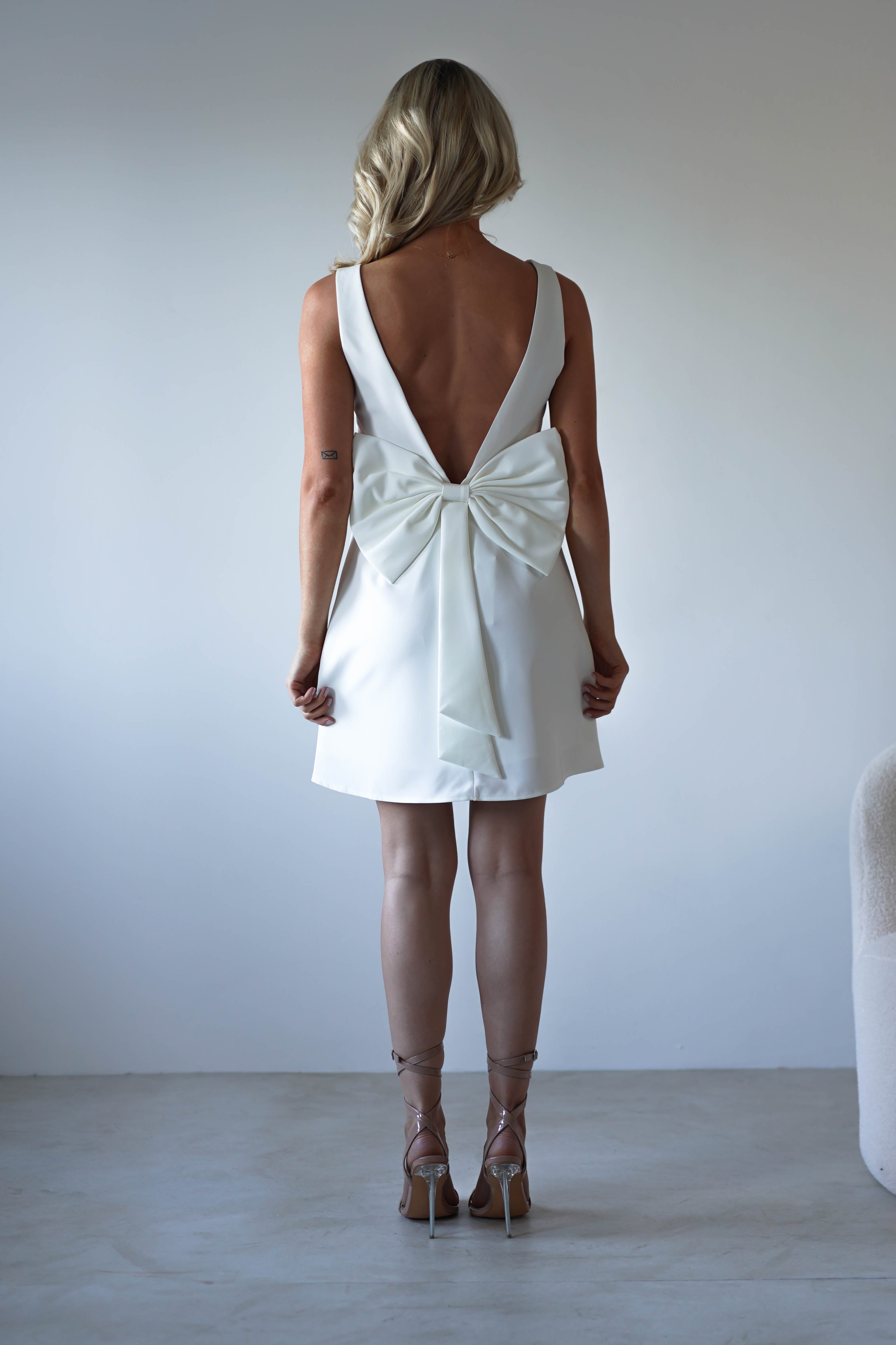 A woman with blonde hair wears a white, sleeveless open back dress with a large bow at the lower back, standing near a light-colored wall. She holds the edges of her party dress and pairs it with strappy high-heeled sandals.