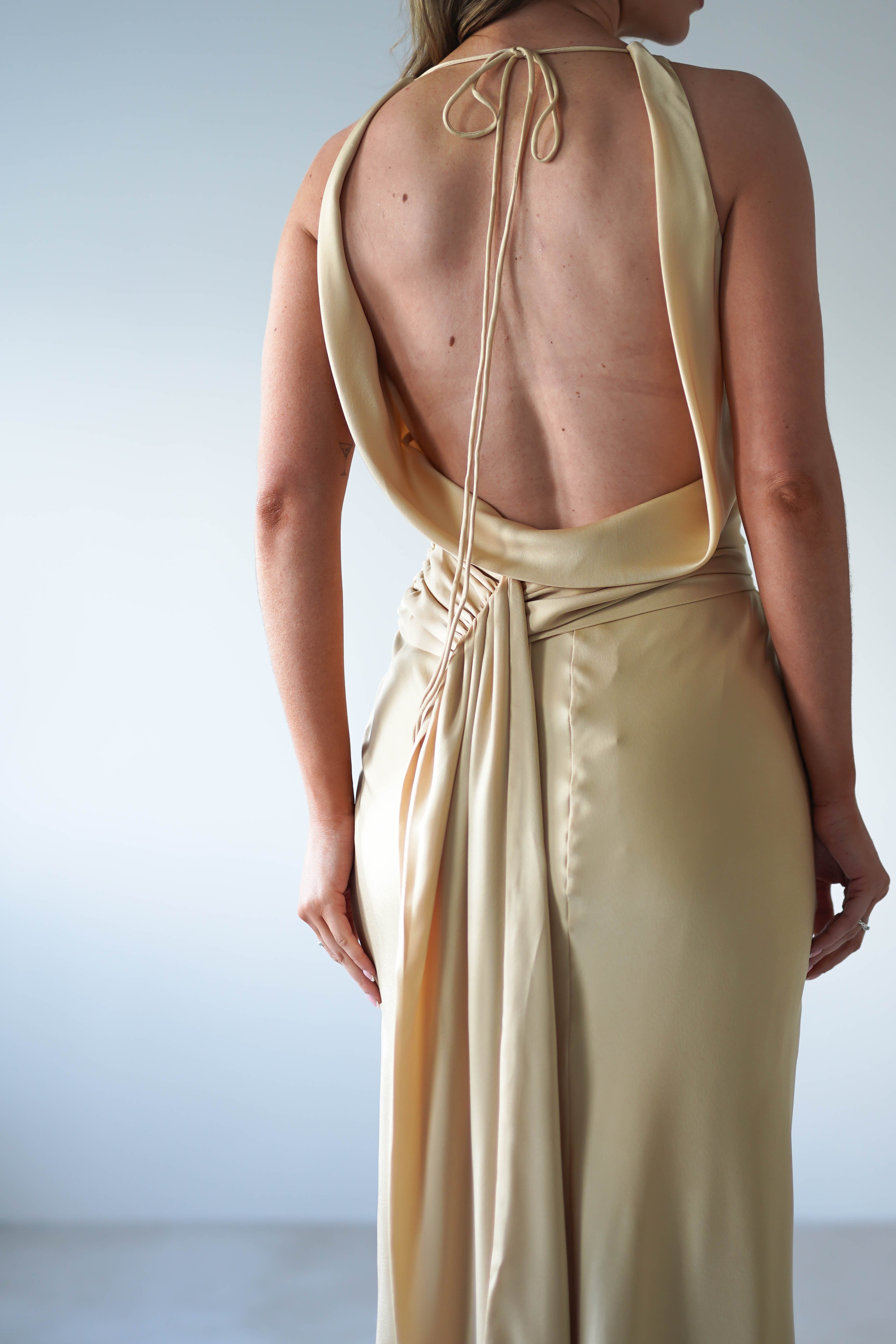 A person wearing a backless, champagne-colored satin maxi dress with a tied halter neck stands against a plain light background. Only their back and part of their arms are visible—an elegant look, perfect for a wedding guest.