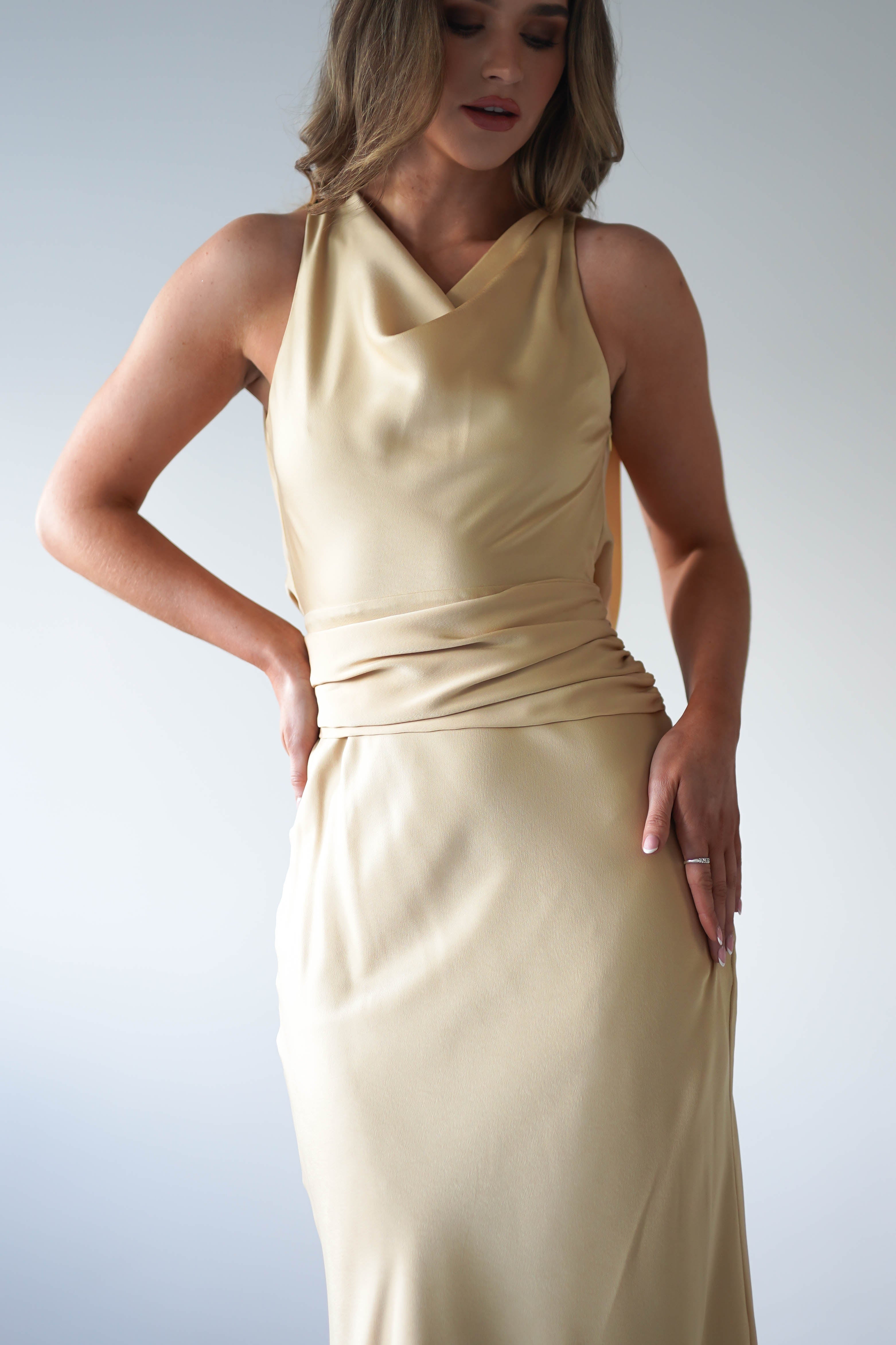 A woman wearing a sleeveless, light beige maxi dress with a cowl neckline and fitted waist stands with one hand on her hip against a plain background—an elegant choice for a wedding guest. Her face is partly visible.