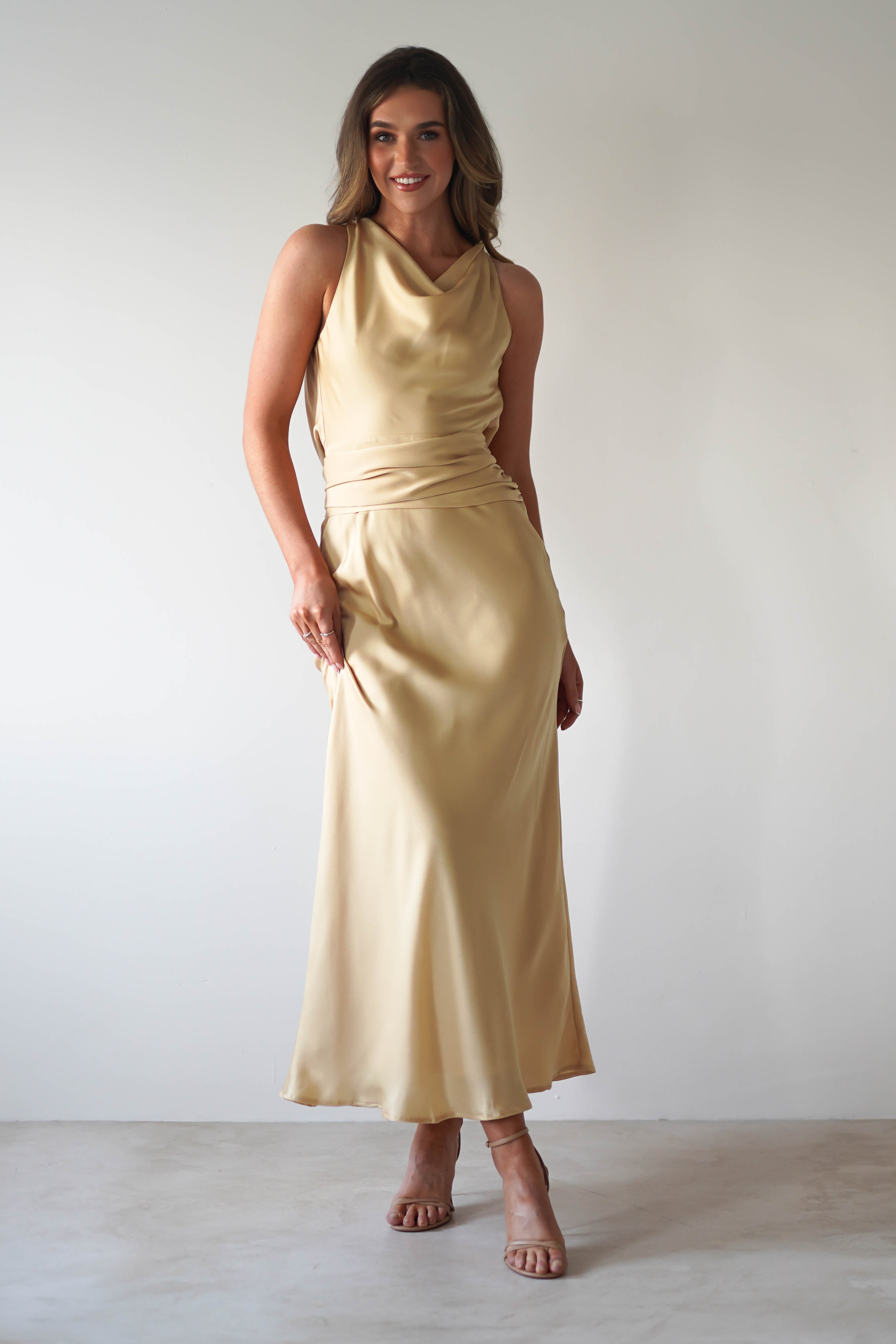 A woman stands against a plain light background wearing a sleeveless, floor-length champagne satin maxi dress with a cowl neckline and draped detailing. She is smiling, with one hand on her hip and the other holding the dress—perfect for a wedding guest.
