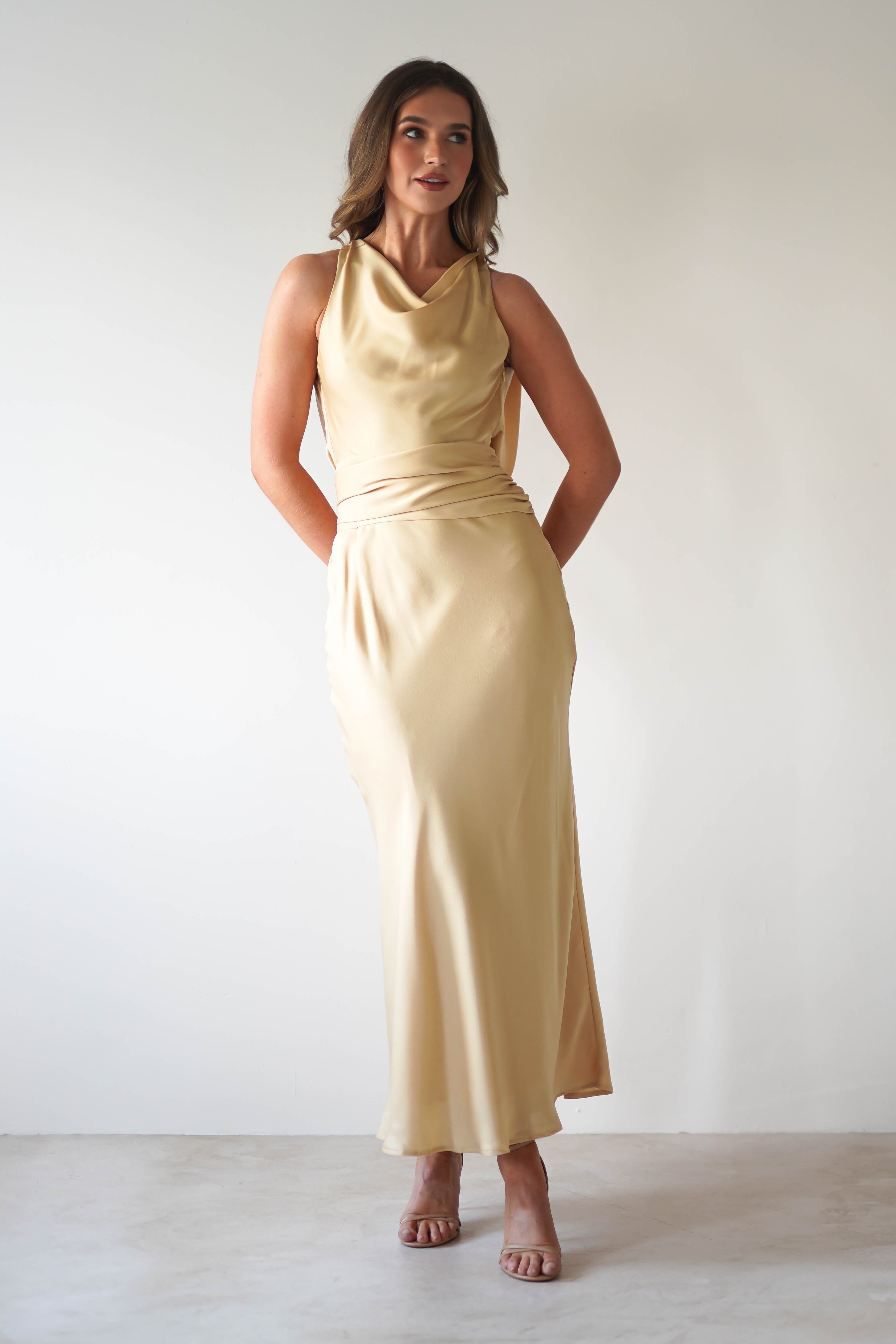 A woman stands against a plain light background wearing a sleeveless, floor-length beige satin maxi dress with a cowl neckline—an elegant choice for a wedding guest. Her hands are behind her back as she gazes slightly up and to the side, in open-toe heels.