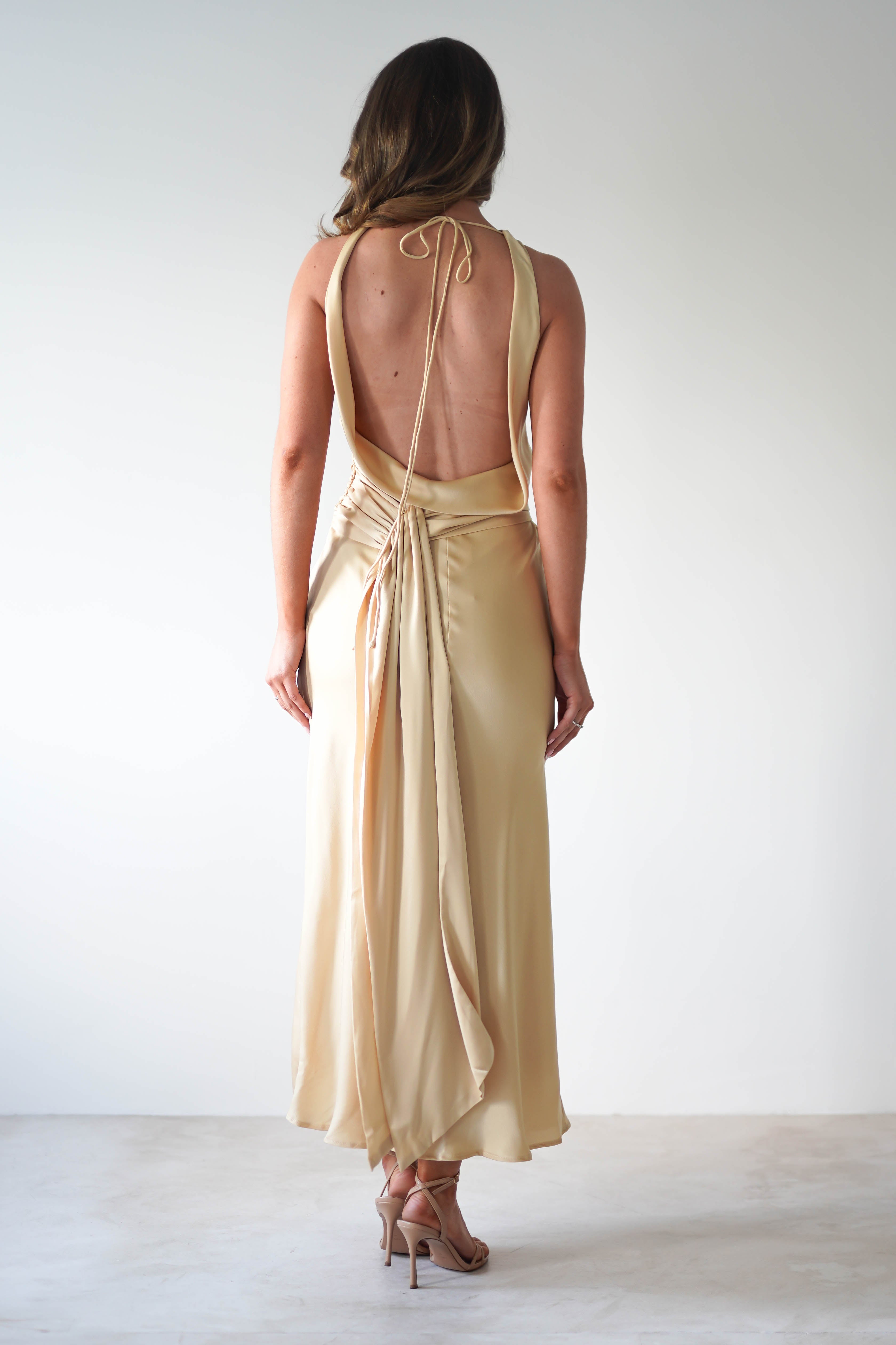 A woman stands facing away, wearing a backless, light gold satin maxi dress with thin straps, a cowl neckline, and a draped tie at the waist—perfect for a wedding guest. She pairs it with nude high-heeled sandals against a softly lit plain background.
