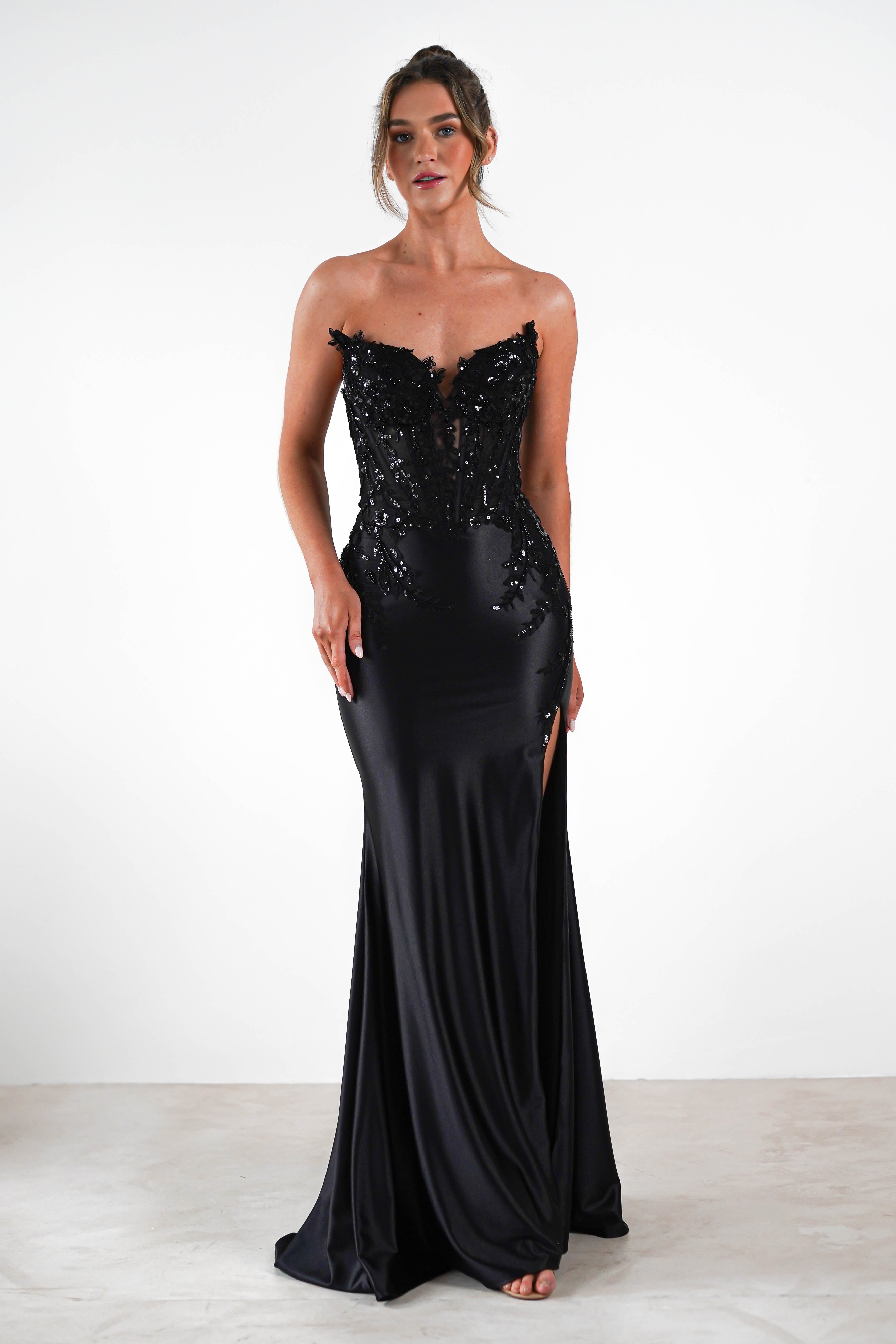 A person in an elegant black evening gown with a sequin embellished bodice stands against a plain white background. Perfect for formal occasions, the dress features a high slit, and the persons hair is styled up.