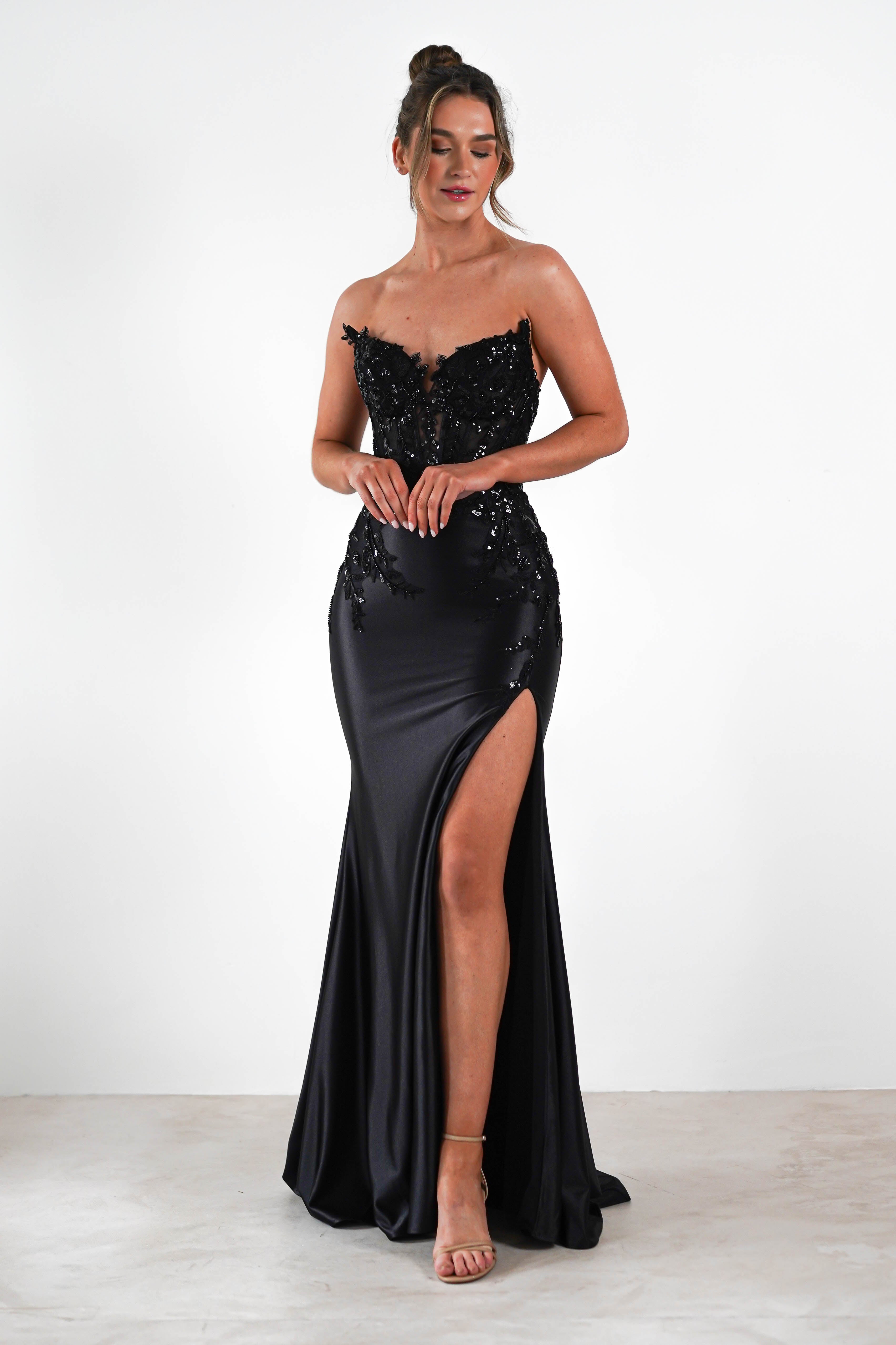 A woman is wearing a strapless black evening gown adorned with intricate lace details and a high slit on one side. She stands against a plain white background, her hair styled in a neat updo, and completes the look with nude strappy heels suited for formal occasions.