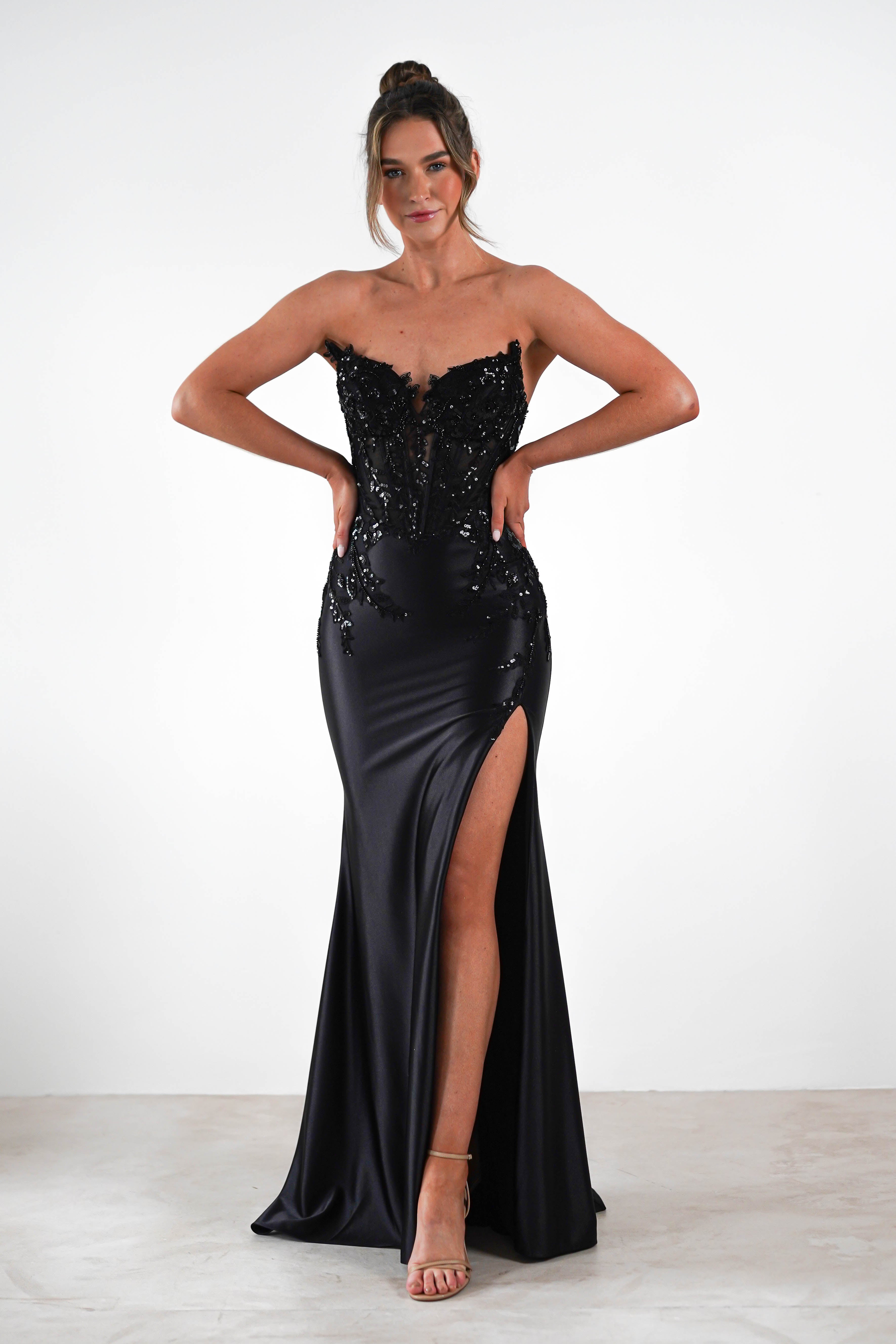 A woman stands confidently in a strapless black evening gown, sequin embellished with a high slit. Her hands rest on her hips, balancing perfectly on high heels. The plain white background accentuates her attire, ideal for formal occasions.