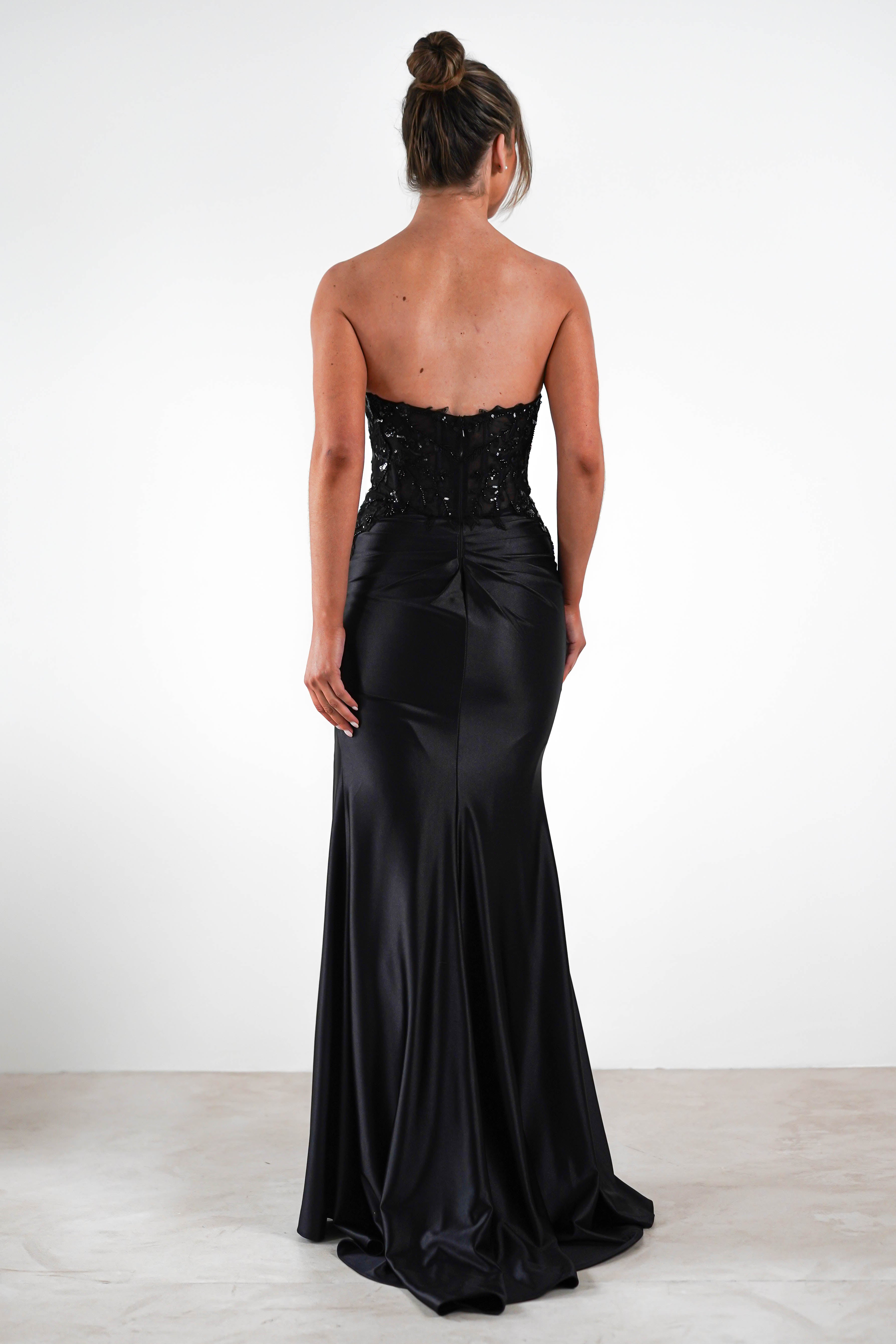 Rear view of a woman in a strapless, floor-length black evening gown with a sequin embellished bodice. Her hair is styled in an updo, perfect for formal occasions, as she stands on a plain floor against a white background.
