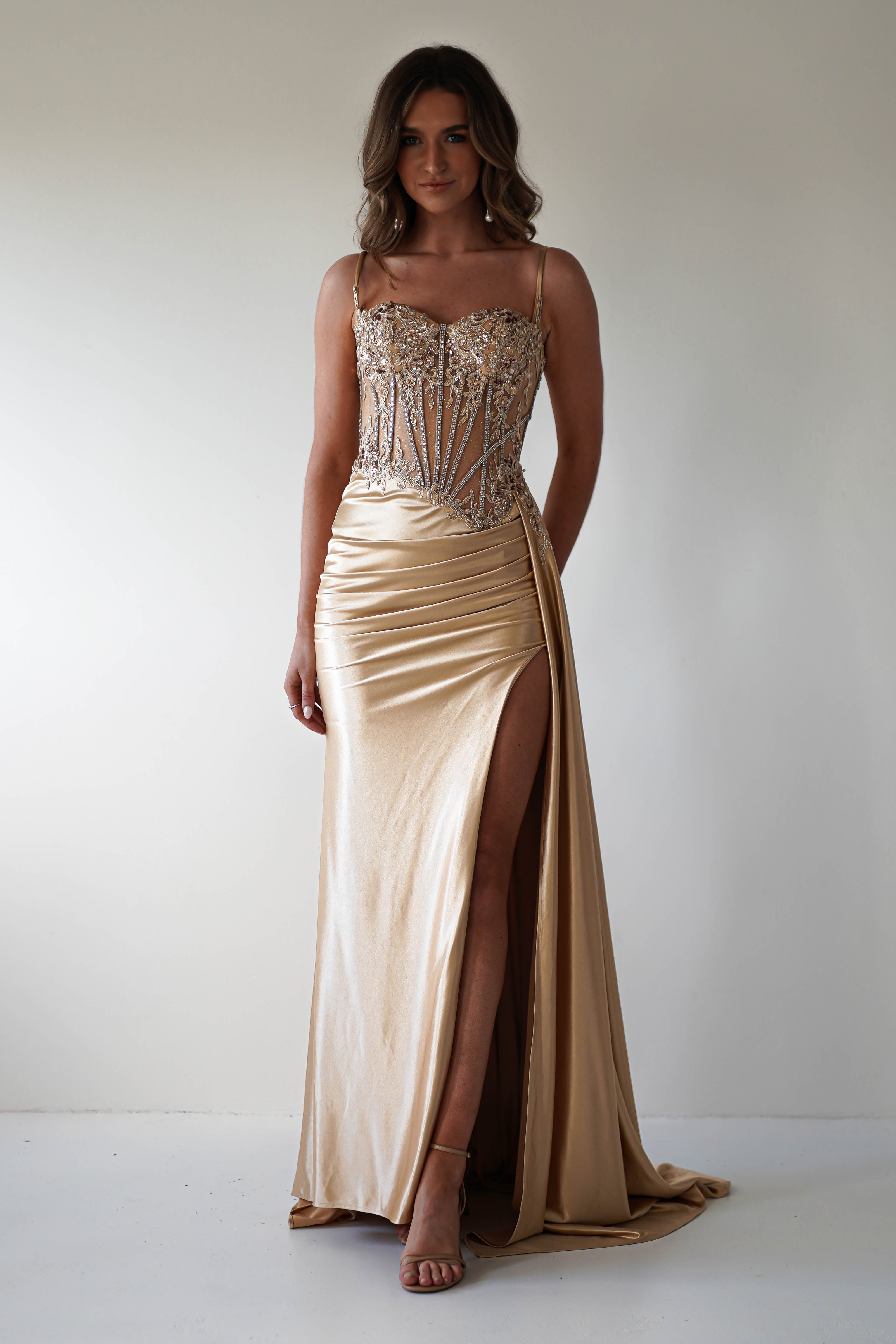A woman stands gracefully in a Diva Embellished Corset Gown, its sleeveless gold fabric shimmering with intricate adornments. The corset bodice accentuates her elegance as she poses confidently in matching heels against a plain white background, with a daring high slit on the skirt.
