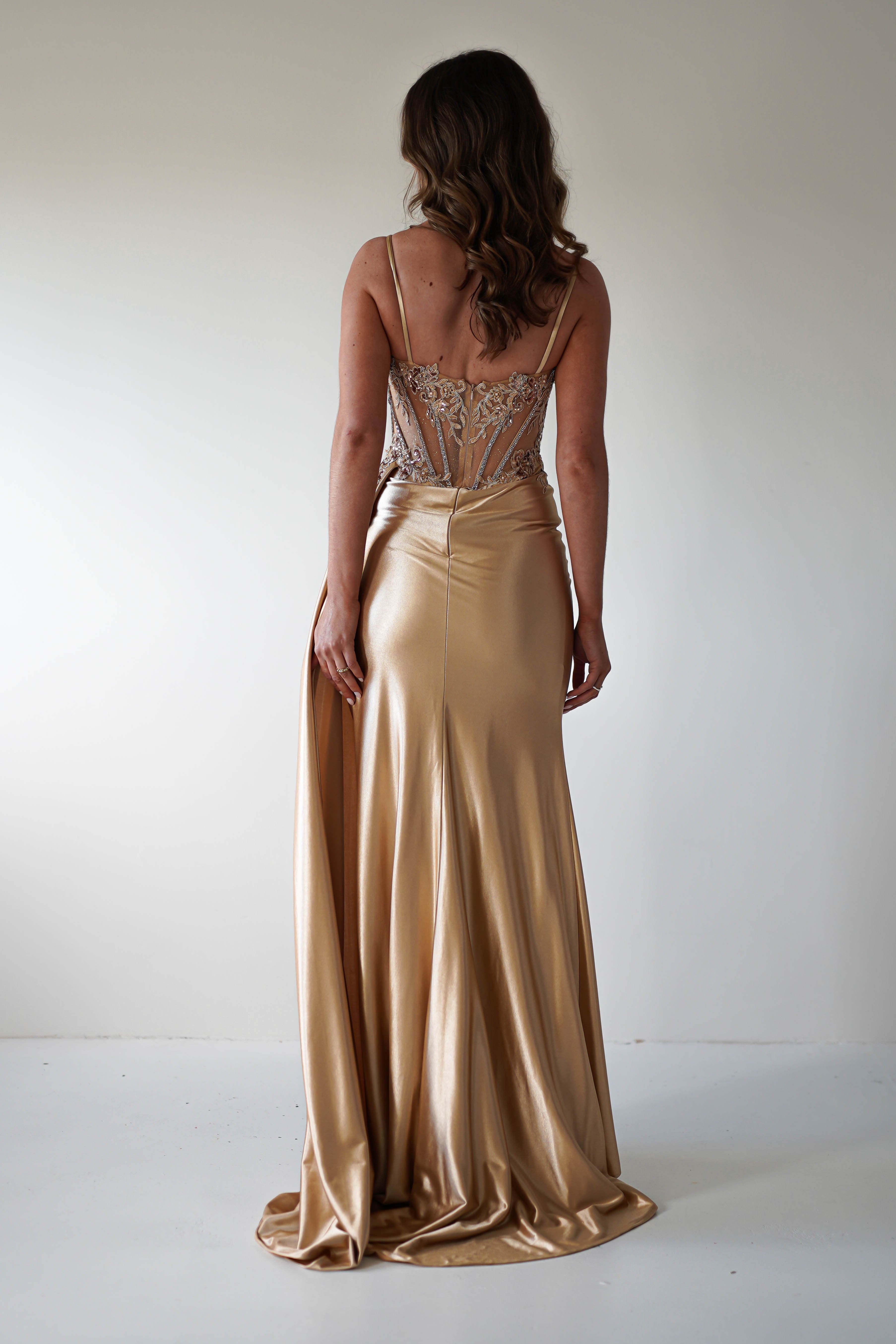 A woman with wavy brown hair stands facing away, wearing a Diva Embellished Corset Gown. The gold, floor-length dress features a fitted bodice with glamorous detailing and thin straps. It flows elegantly, casting a soft silhouette against a plain, light background.