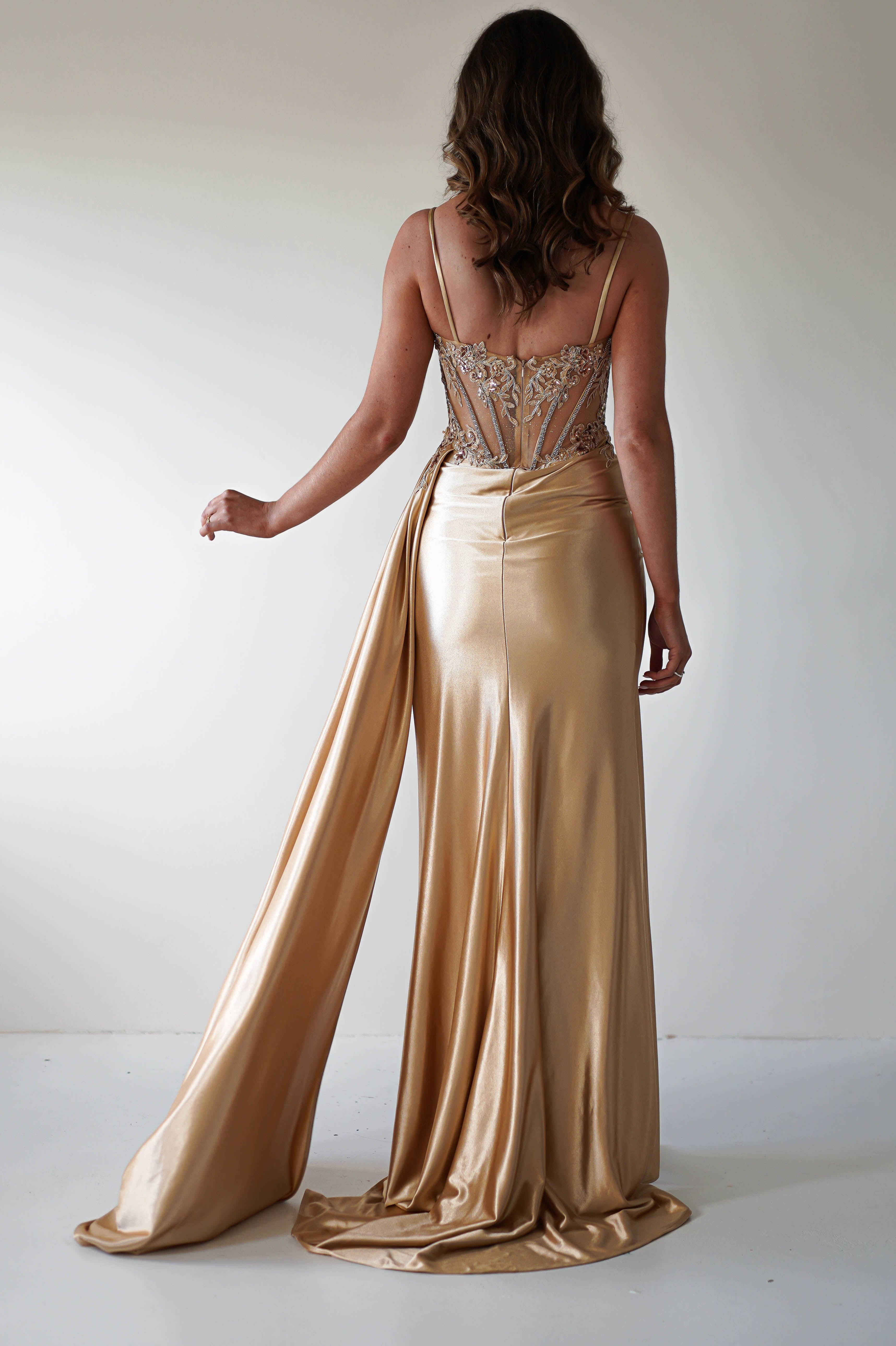 A woman stands with her back to the camera, wearing a golden satin gown with a long train and an embellished bust on a sheer corset bodice. Her wavy hair cascades to her shoulders against a plain white background.