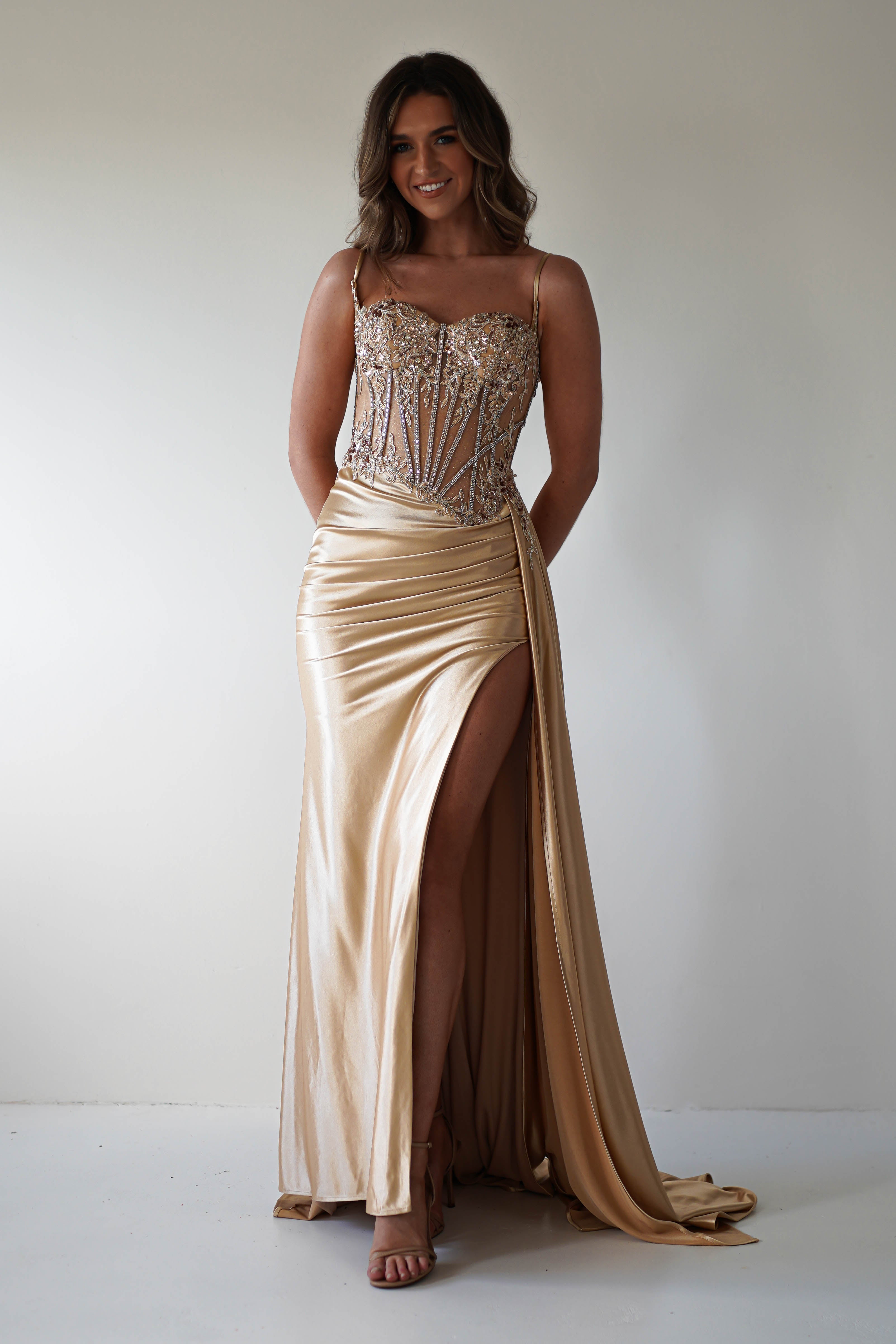 A woman stands smiling, exuding elegance in a shimmering gold Diva Embellished Corset Gown with a high slit and beaded bodice. The gown’s thin straps and flowing skirt complement her long, wavy brown hair as she poses against a plain background.