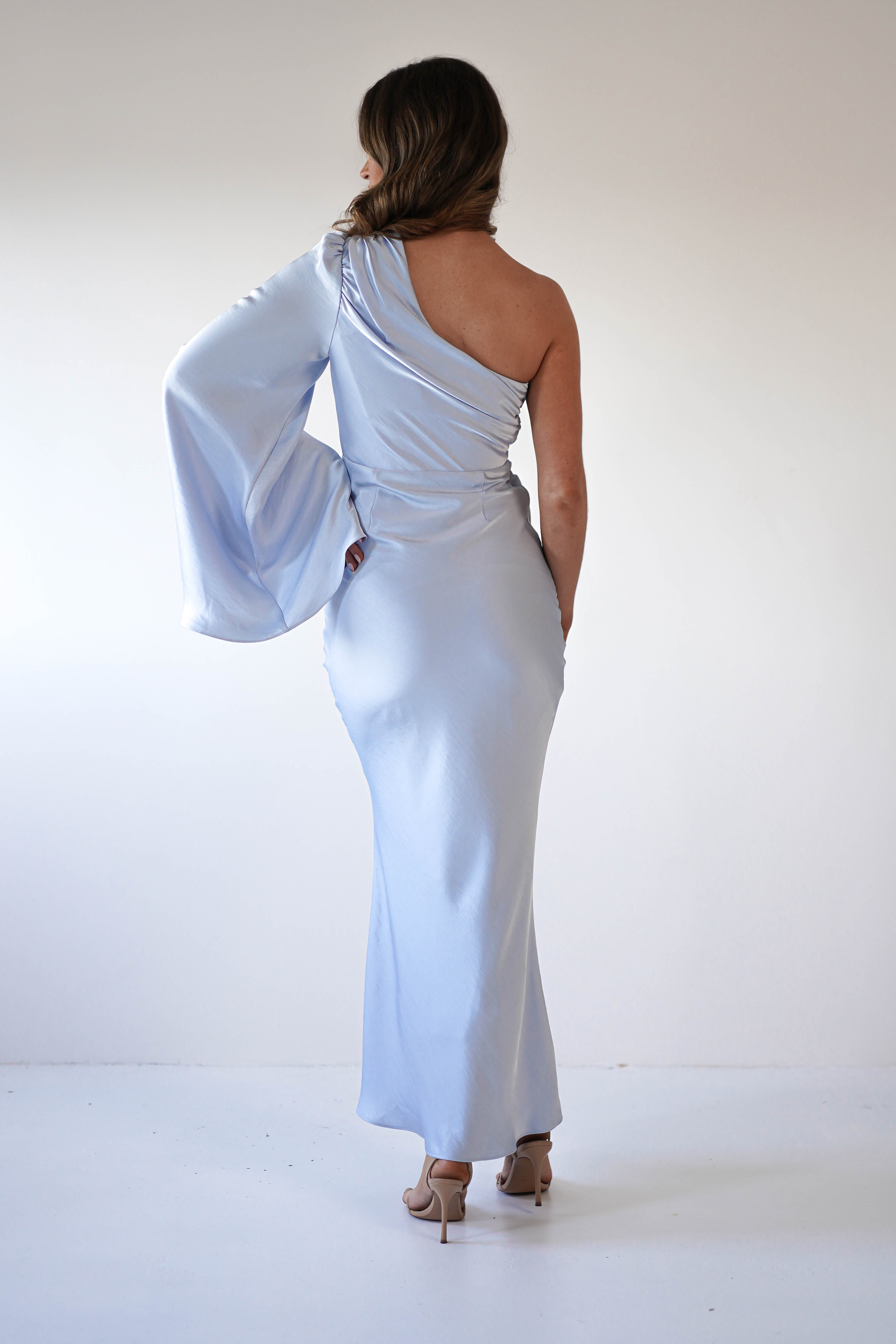 A woman stands facing away, wearing a silky fabric, light blue maxi dress with an asymmetrical neckline, draped sleeve, and fitted silhouette. She has wavy brown hair and nude heels, set against a plain white background.