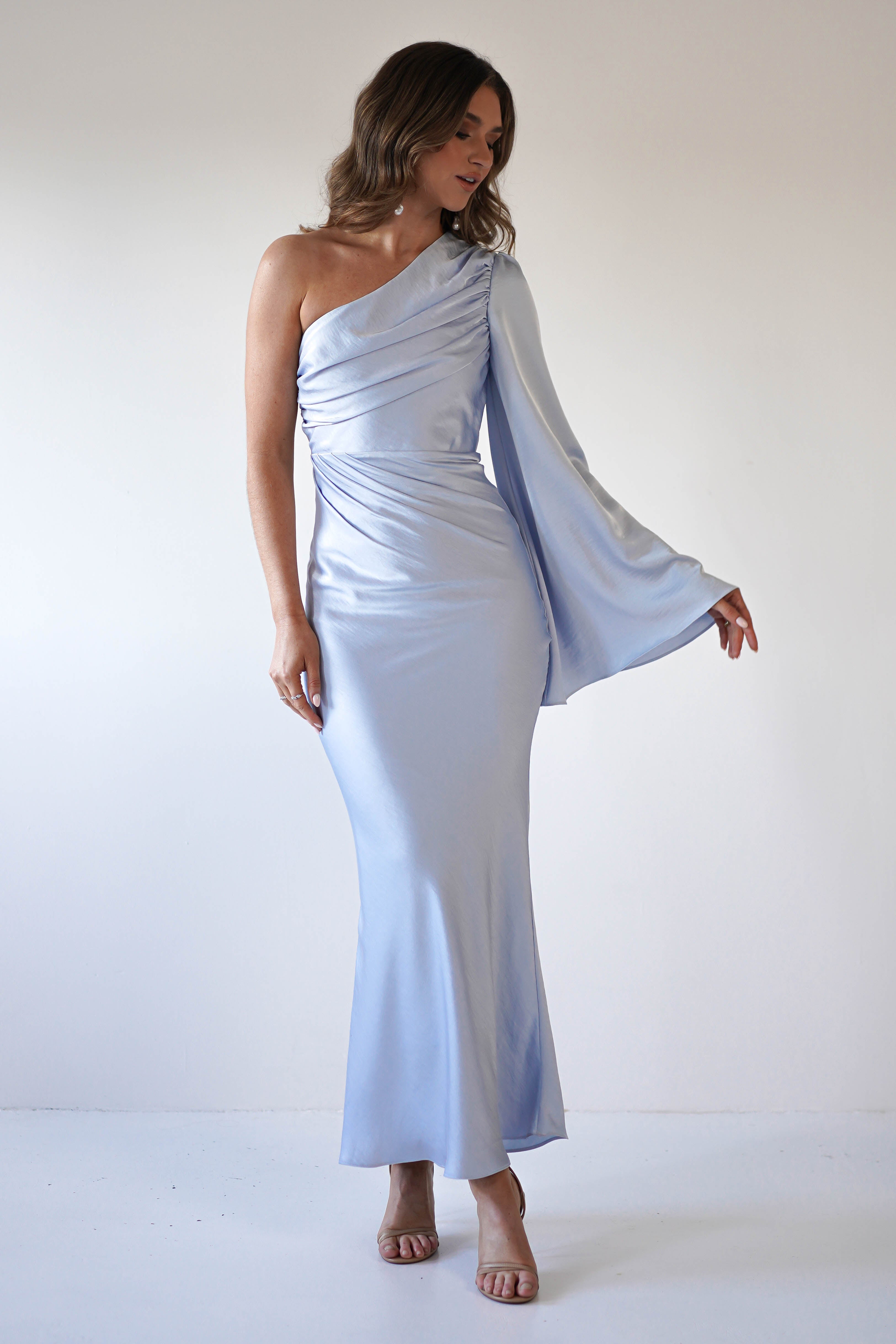 A woman wearing a light blue, one-shoulder maxi dress with an asymmetrical neckline, draped bodice, and single long, flowing sleeve crafted from silky fabric stands against a plain white background.