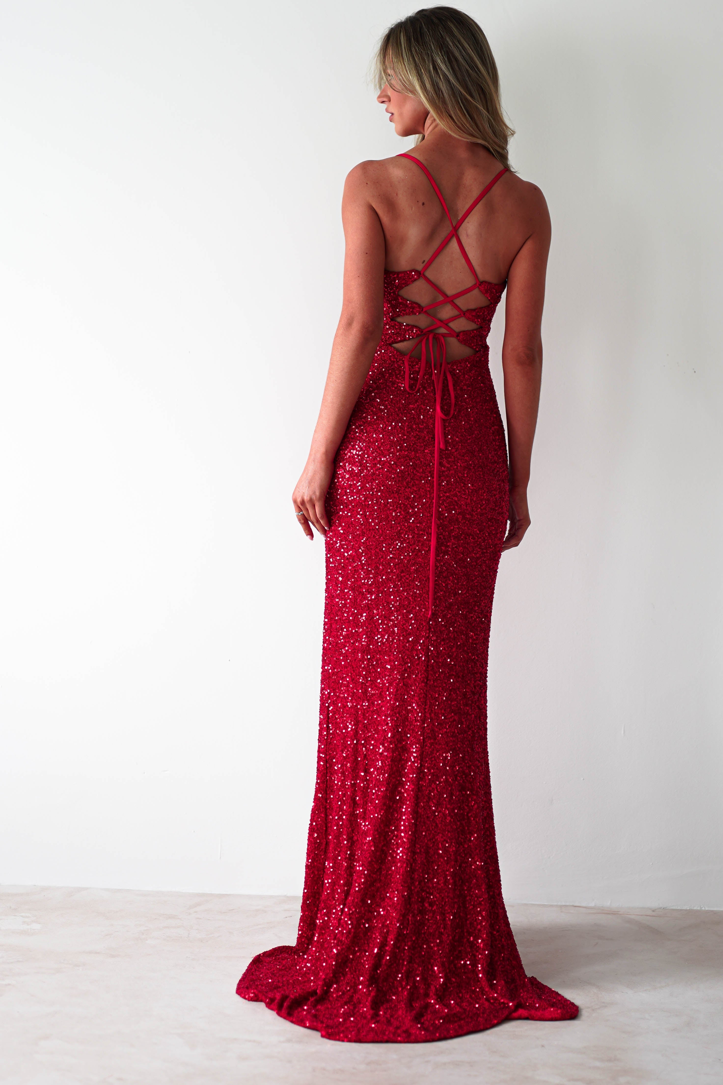 A woman in a sparkling red sequin mermaid maxi stands with her back to the camera. The polyester dress features a lace-up back and floor-length hem. Her long blonde hair cascades down as she poses against a plain, light-colored background.