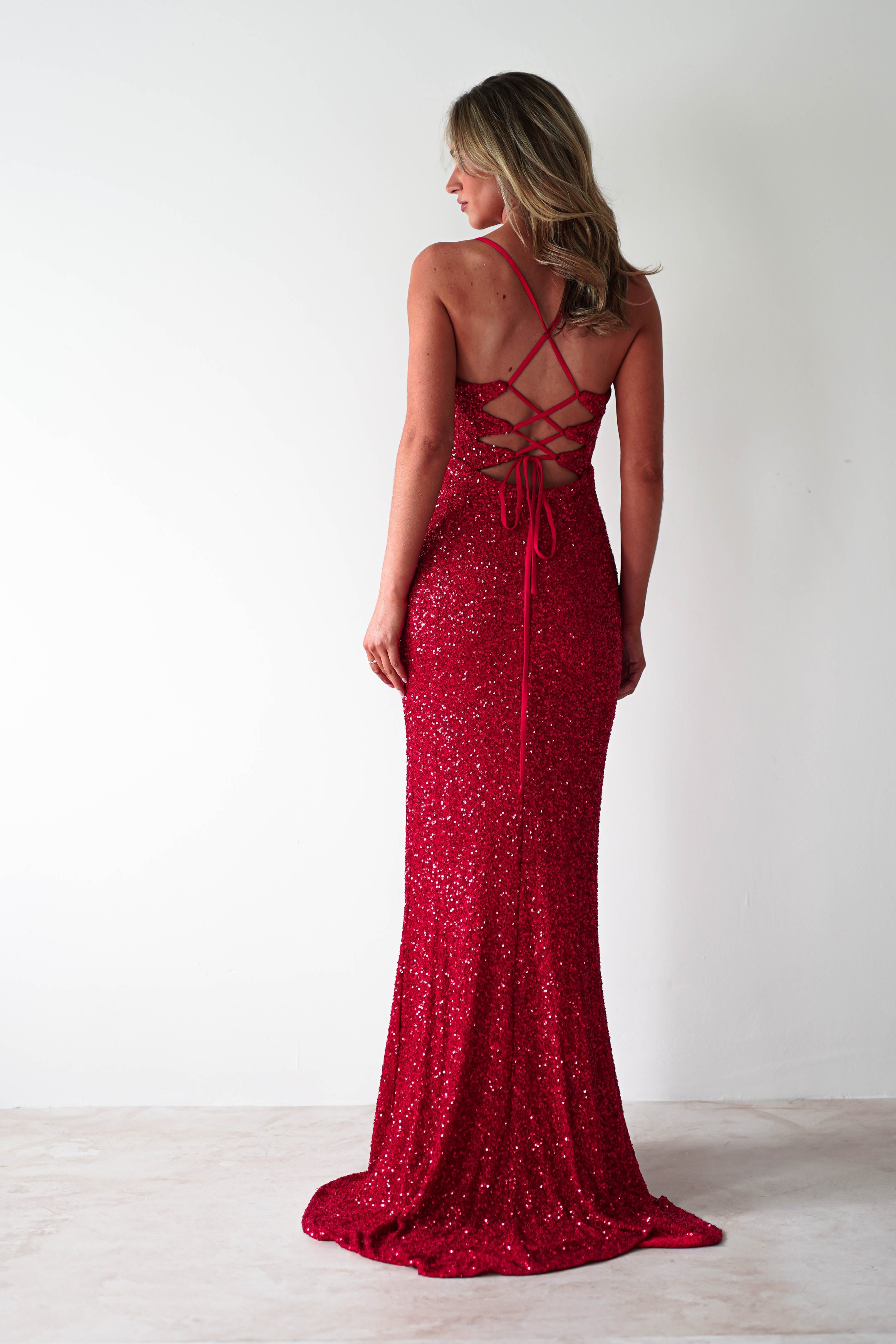 A woman in a red sequin mermaid maxi with a crisscross lace-up back stands on a light floor against a white wall. She is facing away, showcasing the elegant design of her polyester dress.