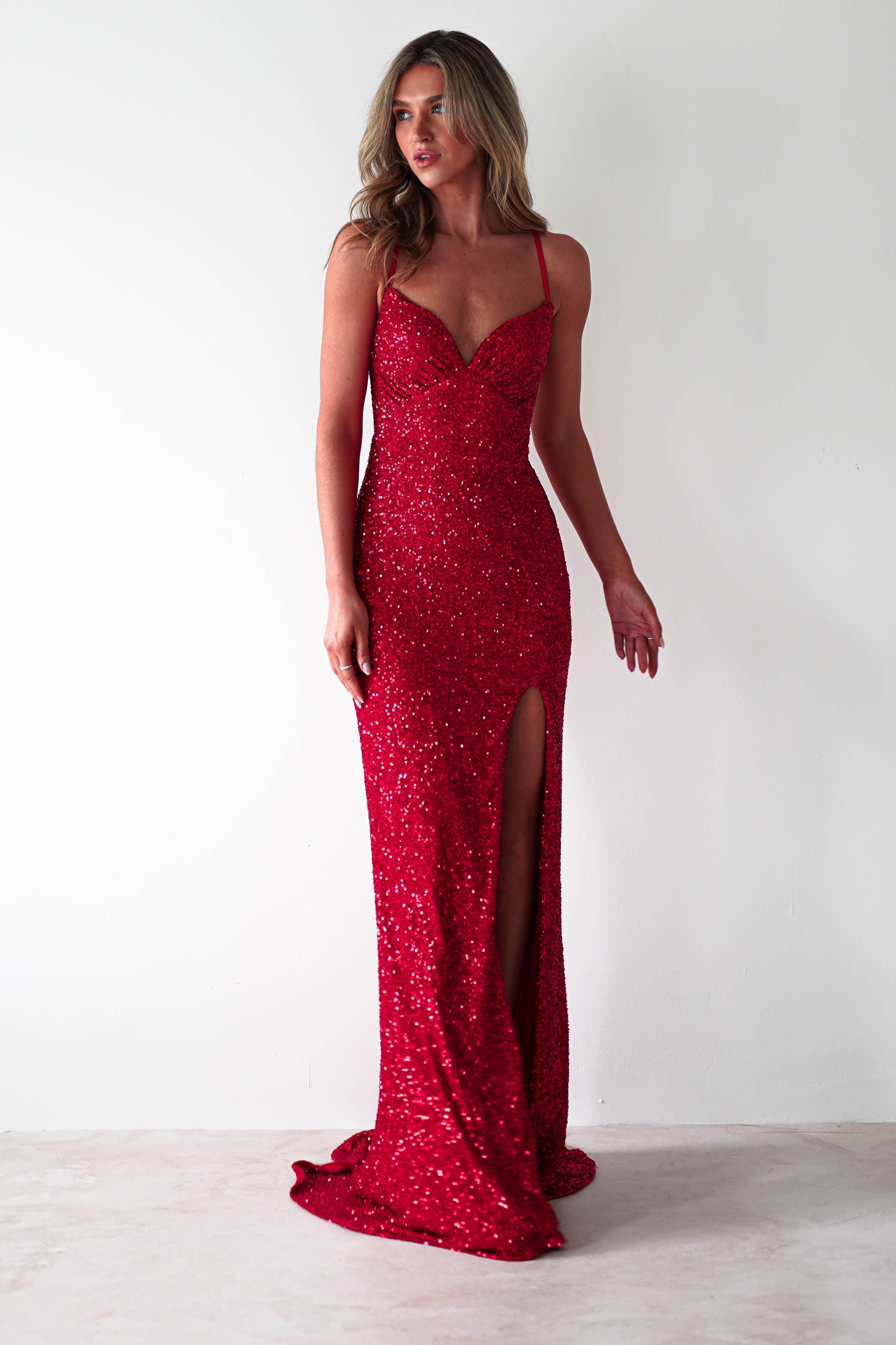 A woman in a sparkling sequin mermaid maxi with thin straps and a lace-up back stands against a light background, looking to the side. The polyester dress is form-fitting and features a high slit, elegantly extending to the floor.