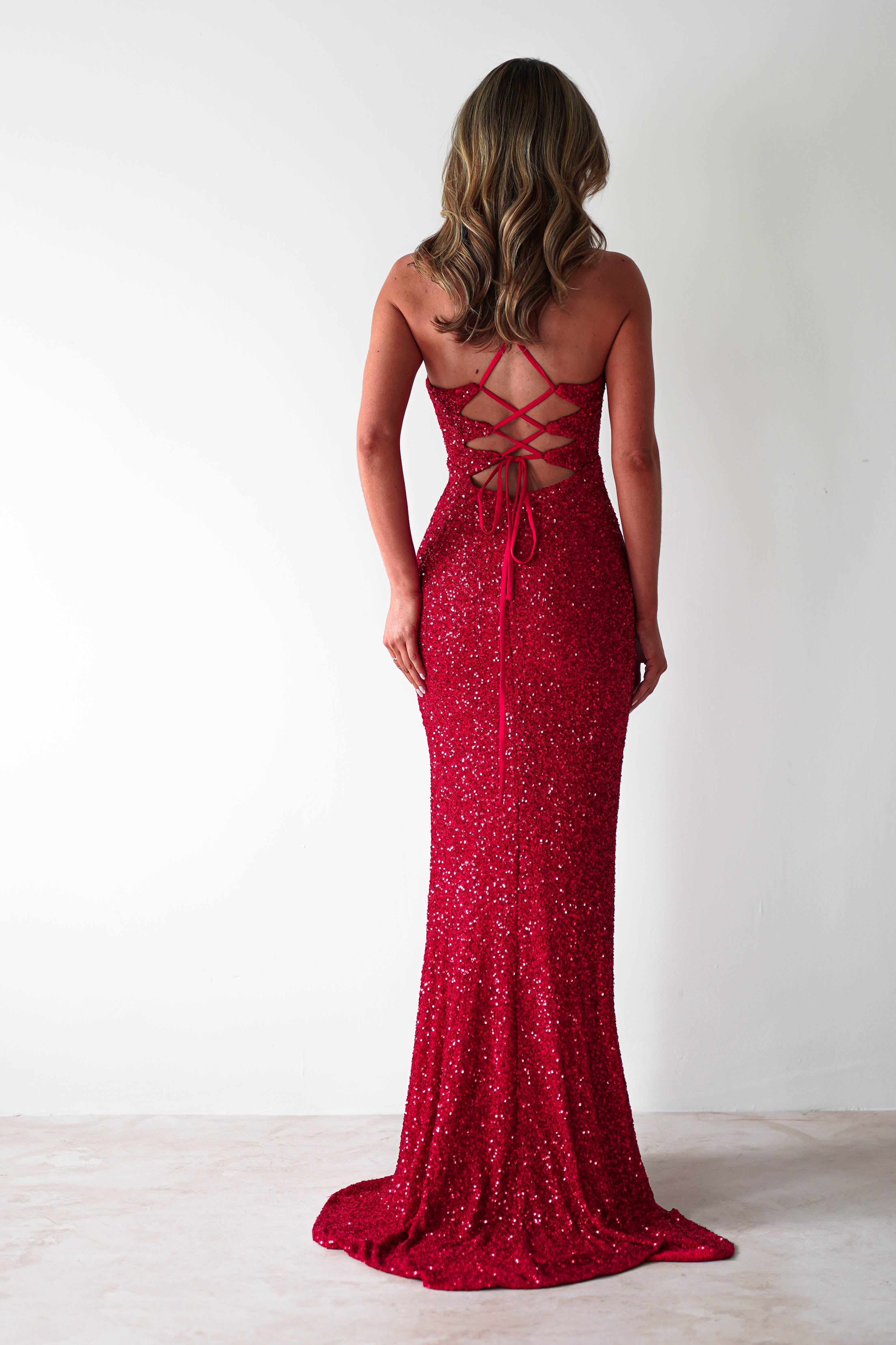 A woman with wavy hair stands facing a white wall, wearing an elegant sequin mermaid maxi gown. The backless design features a crisscross lace-up detail, and the dress hugs her figure before cascading to the floor.