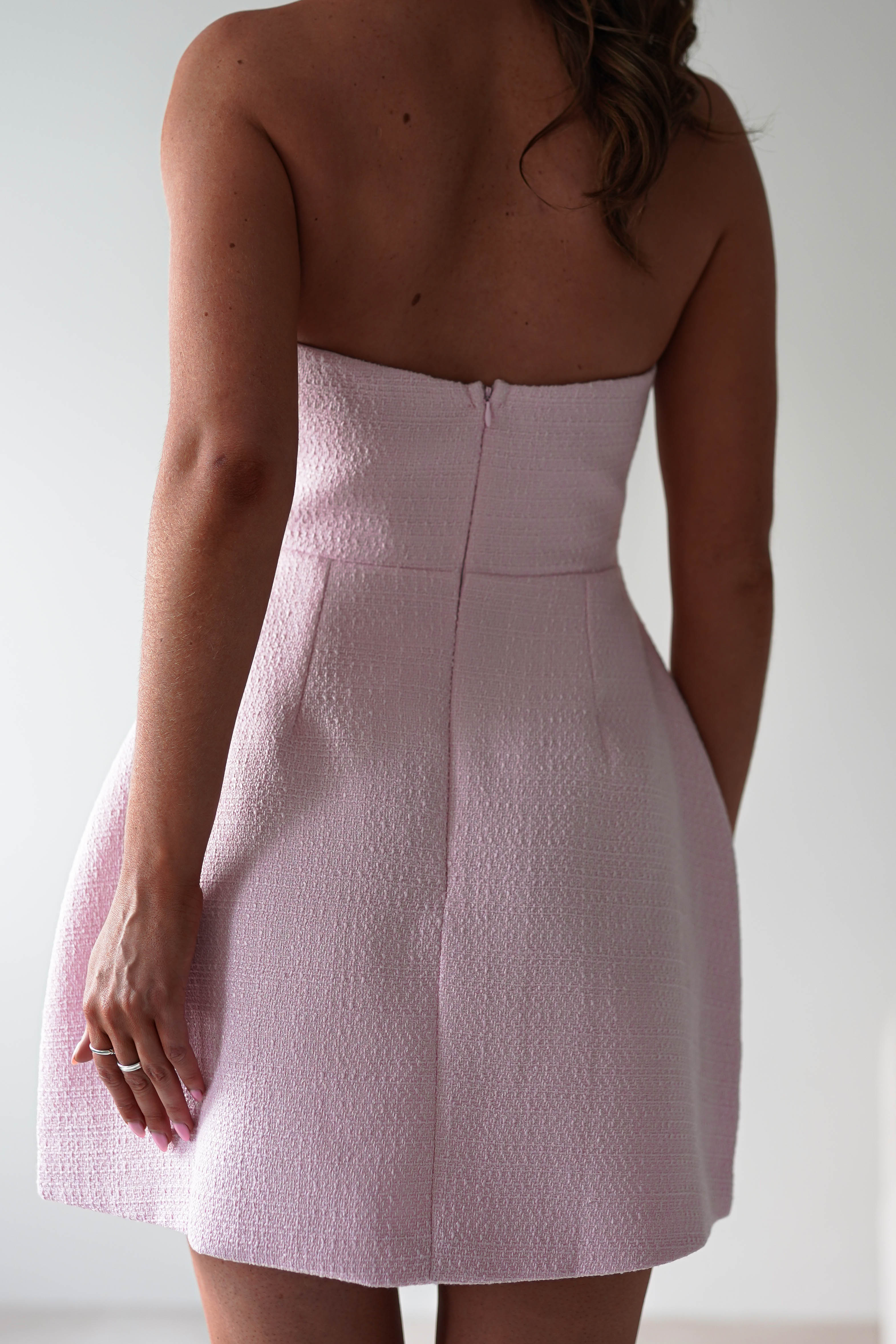 A woman stands with her back to the camera in a light pink tweed textured dress, perfect for a wedding guest. Her left arm is bent as she holds her right hand by her side against a plain, light background.