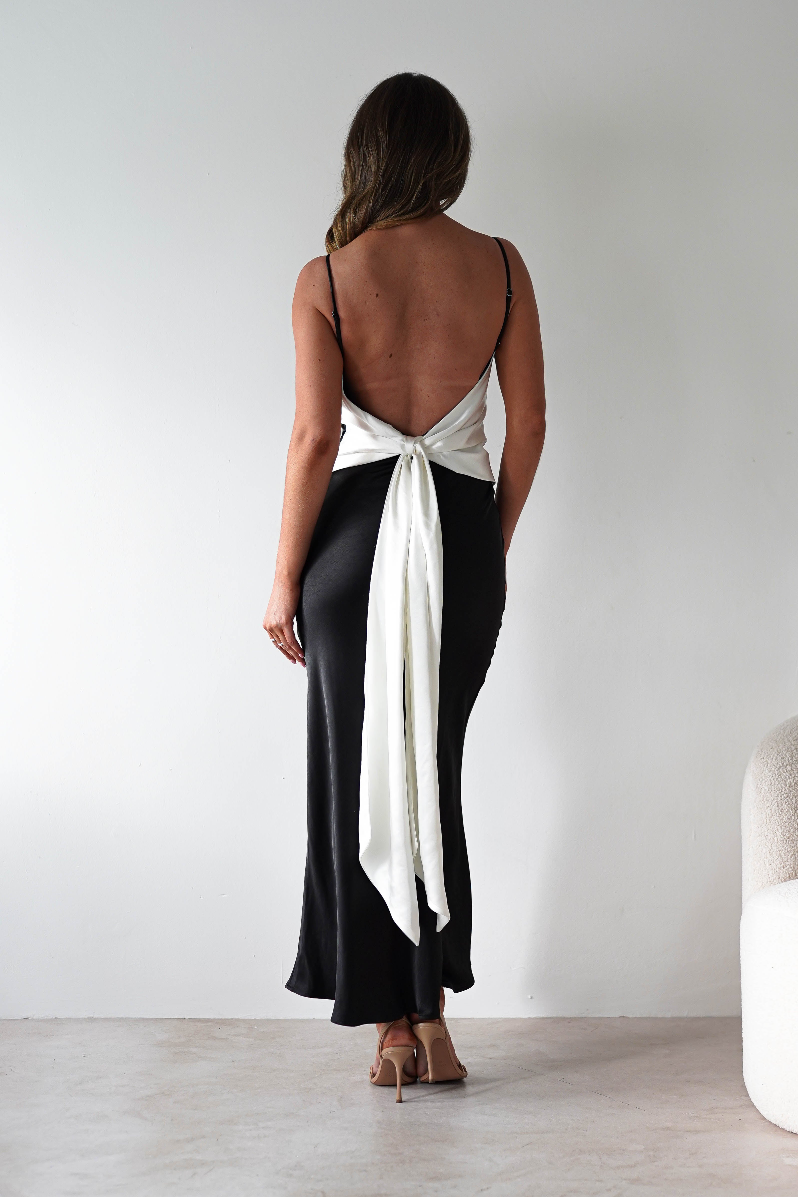 A woman stands facing away in a satin black midi dress with a back tie detail—a large white bow at the waist. Her long, wavy brown hair and beige high-heeled sandals complete this chic look, ideal for bridesmaids, against a plain white wall.