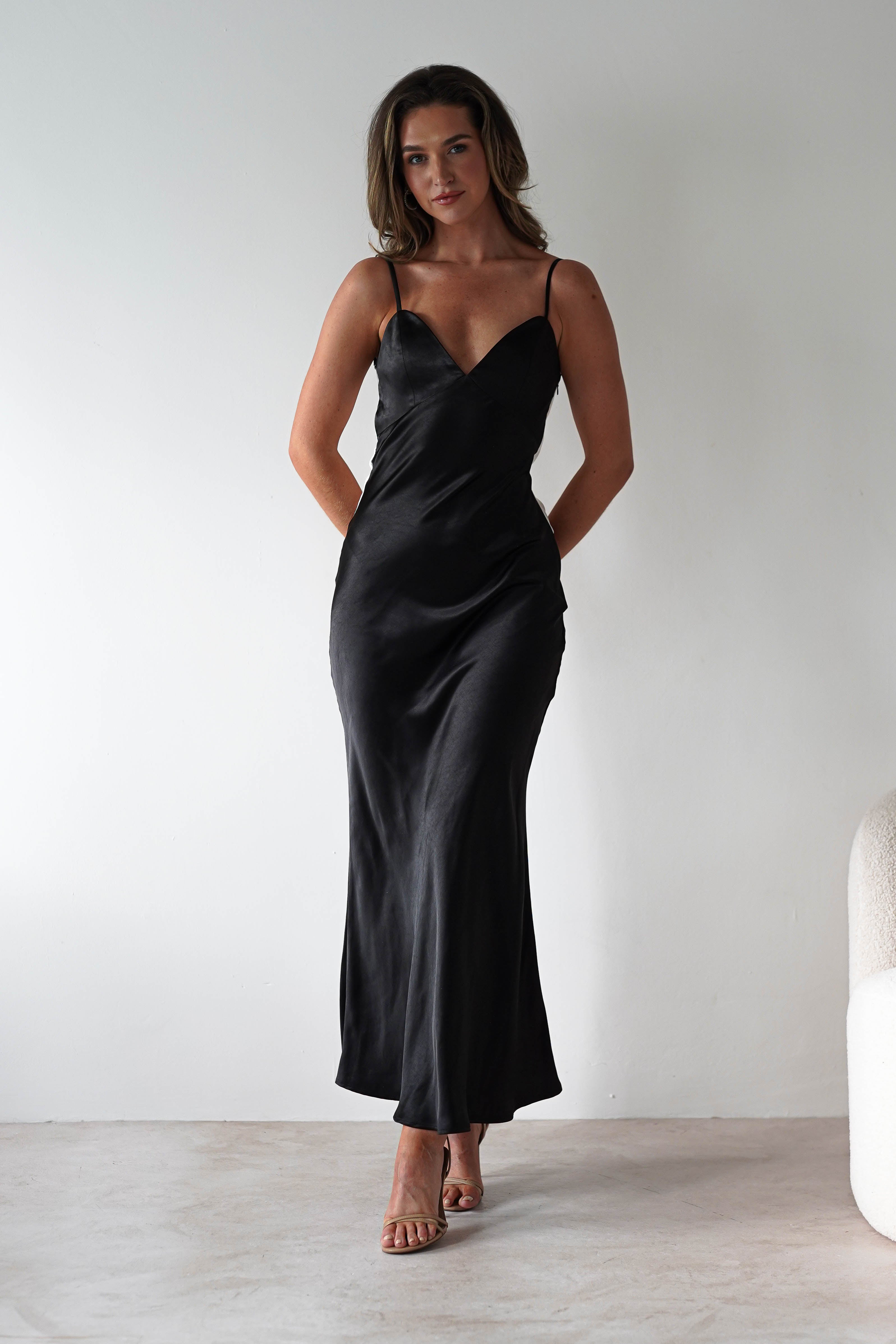A woman stands against a plain white wall, wearing a satin black midi dress with thin straps and back tie detail, paired with nude heeled sandals. With her hands behind her, she looks confidently at the camera—a modern choice for bridesmaids.