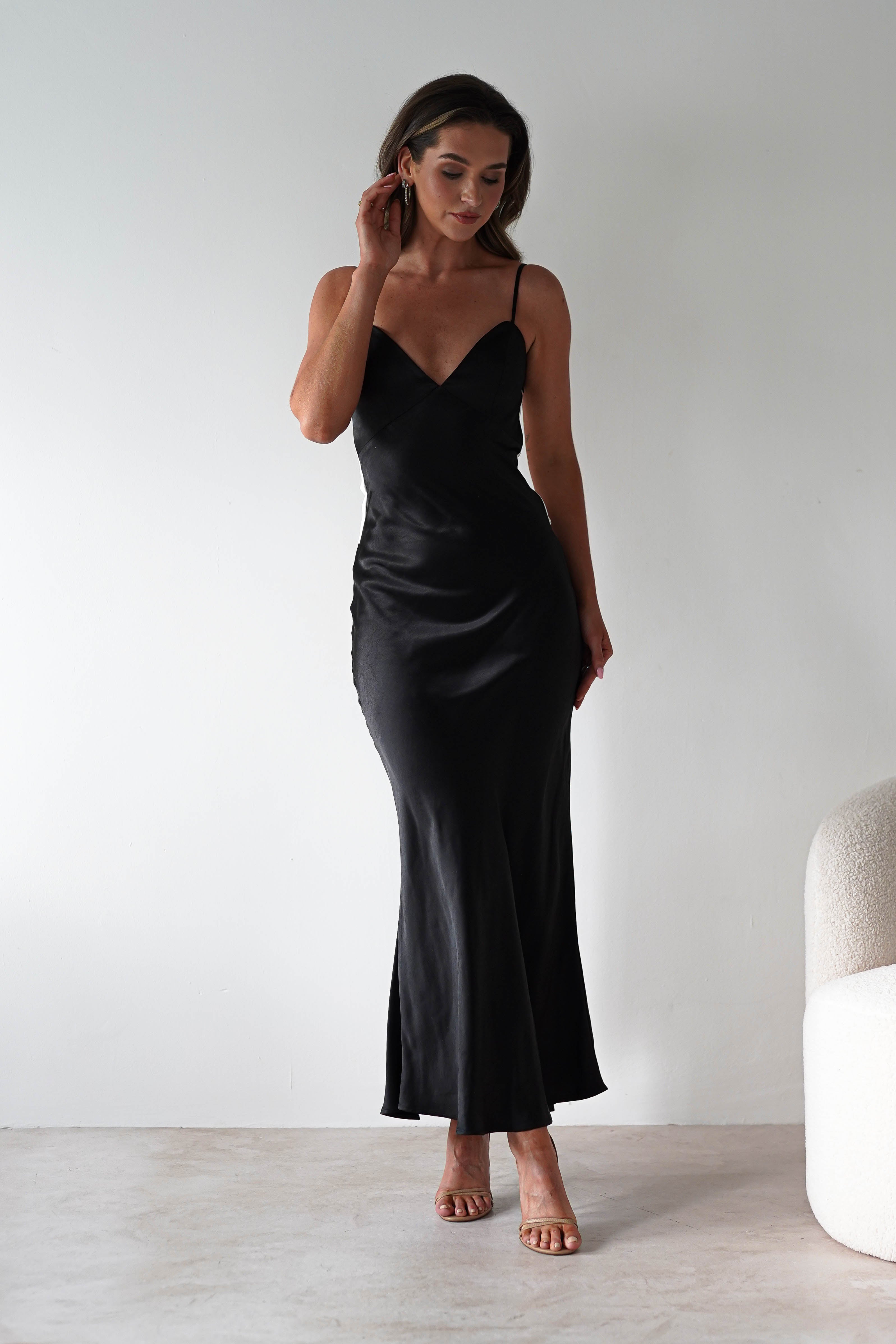 A woman in a sleeveless satin black midi dress with back tie detail and nude heels stands indoors against a plain white wall, gently touching her hair, with a light-colored armchair partially visible on the right. Perfect for bridesmaids or special events.