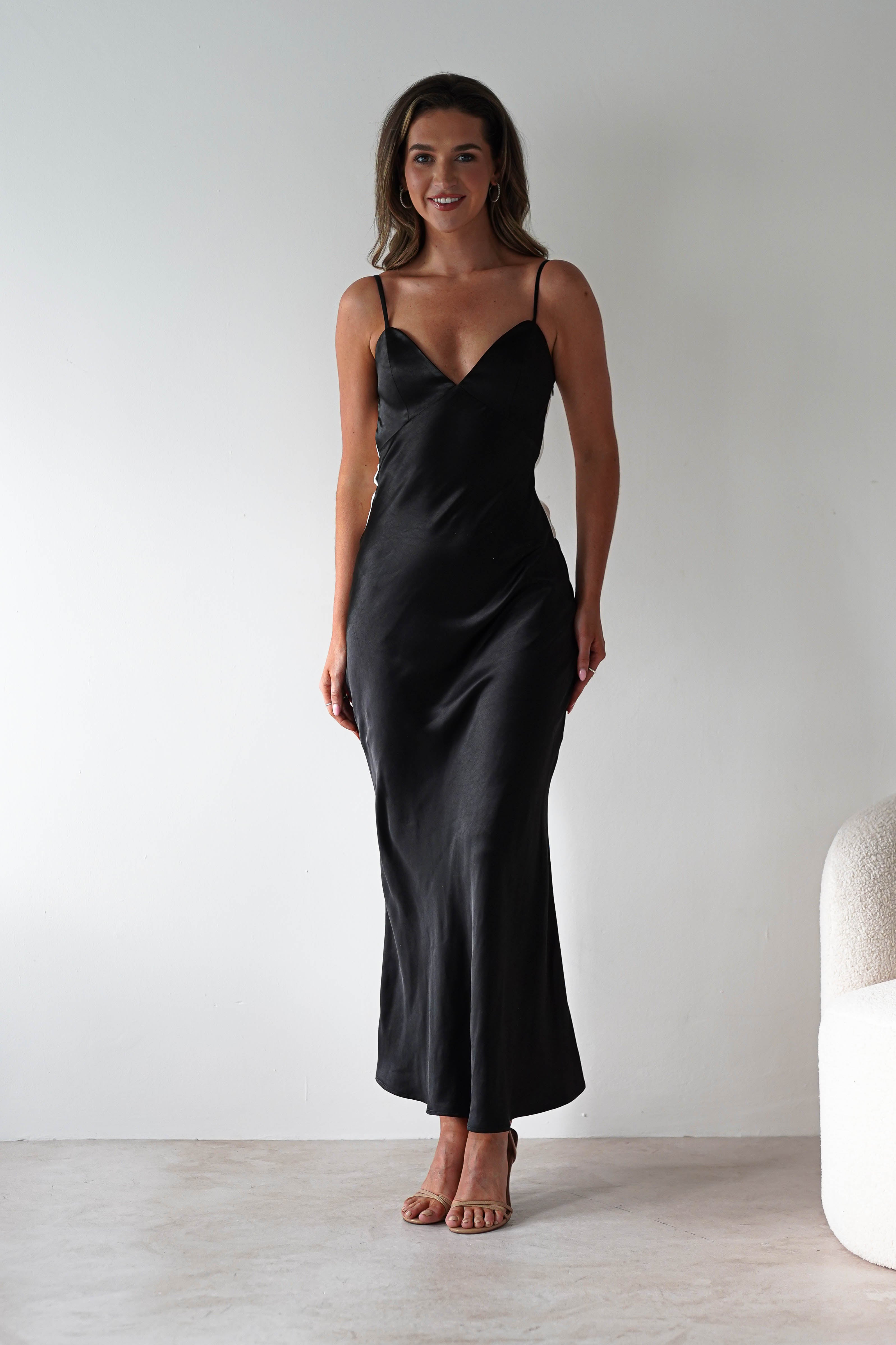 A woman stands against a plain white wall, wearing a satin black midi dress with thin straps and nude heels. The elegant look, perfect for bridesmaids, is completed by her long brown hair and a smile. Part of a white textured chair appears on the right.