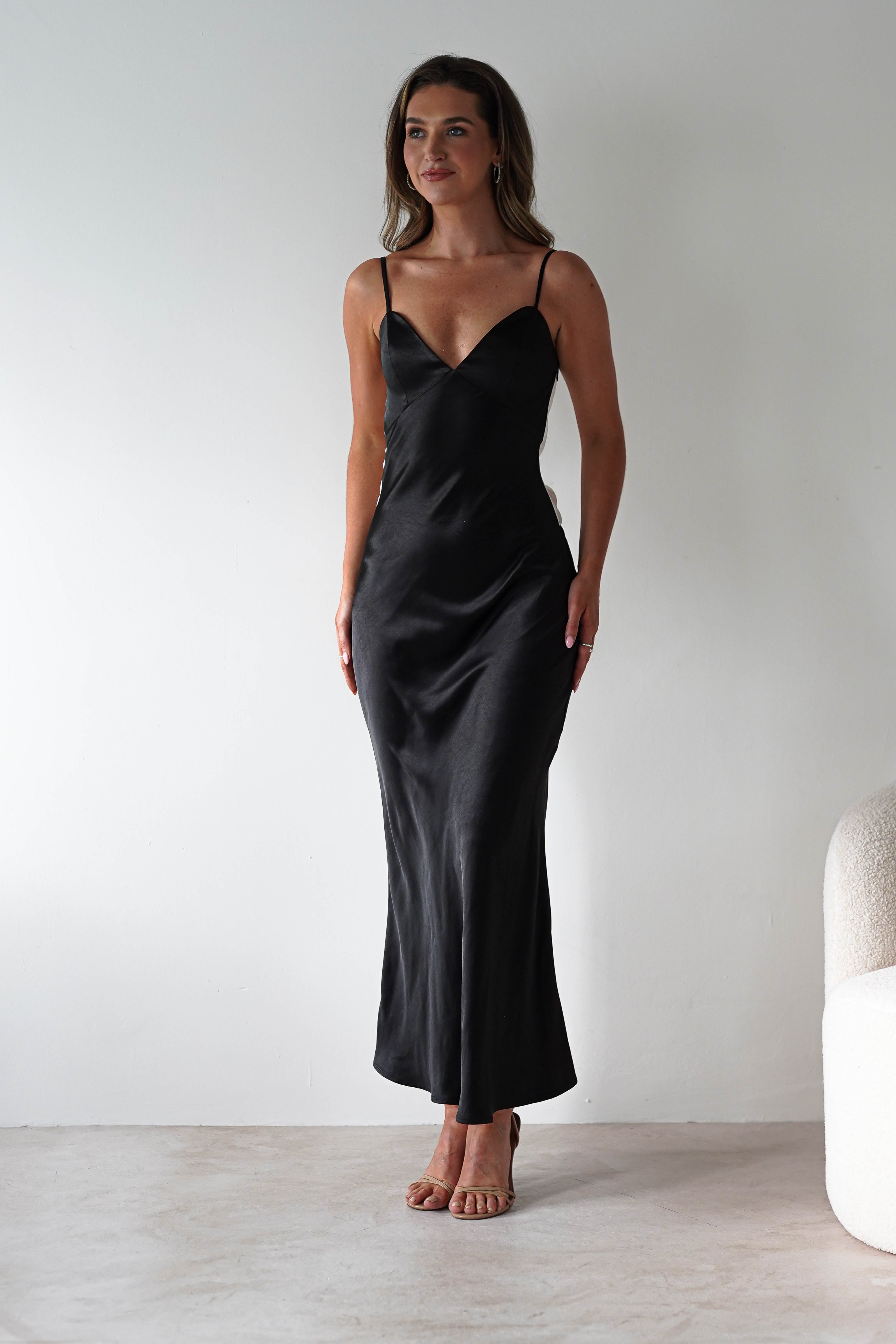 A woman stands against a plain white wall, wearing a satin black midi dress with thin straps and a back tie detail—perfect for bridesmaids. Her hands rest gently at her sides, and she has long brown hair and a slight smile.