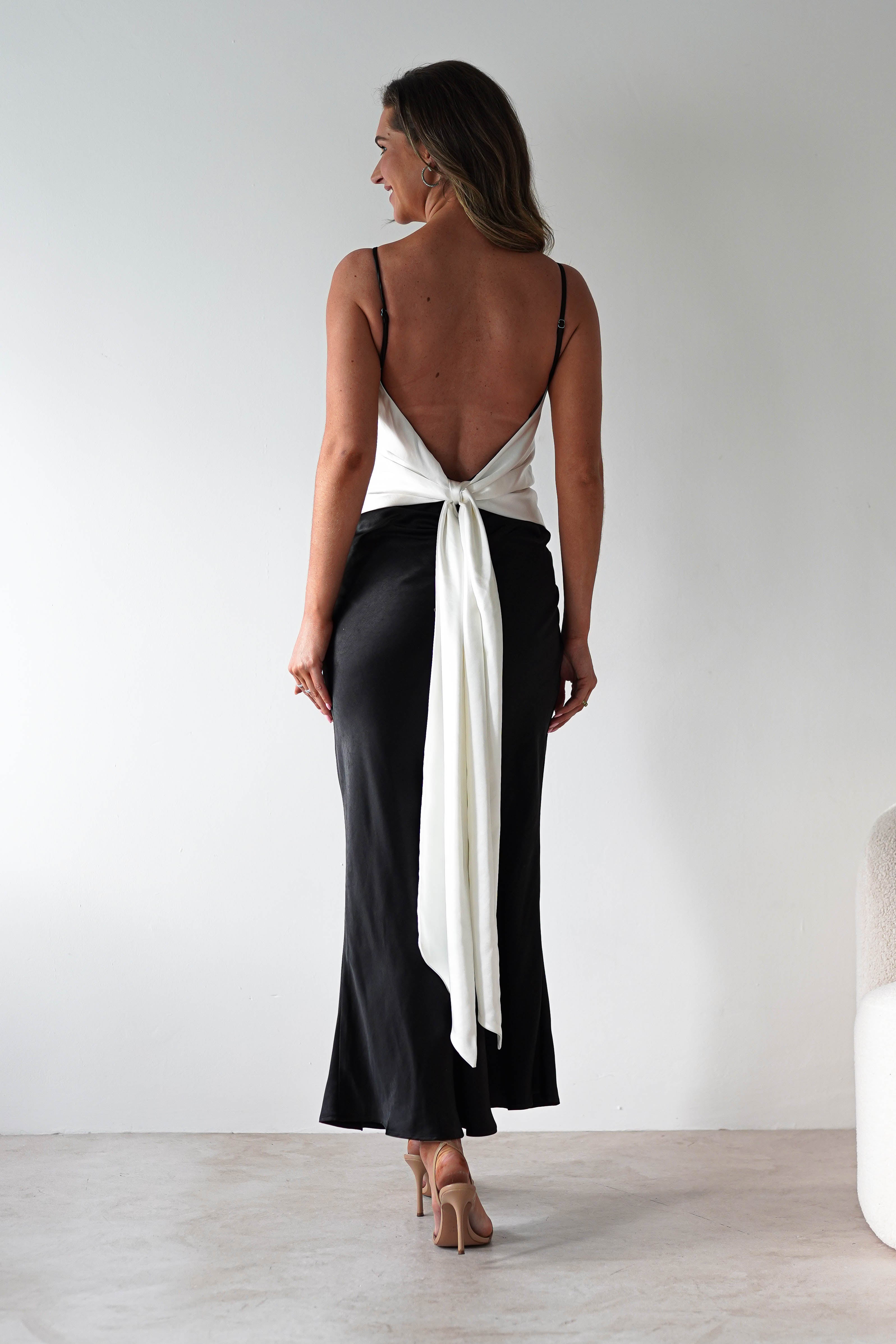 A woman stands facing away, wearing a satin black midi dress with a deep open back and elegant back tie detail. She has wavy brown hair, nude heels, and is poised in a minimal, light room—perfect inspiration for bridesmaids.