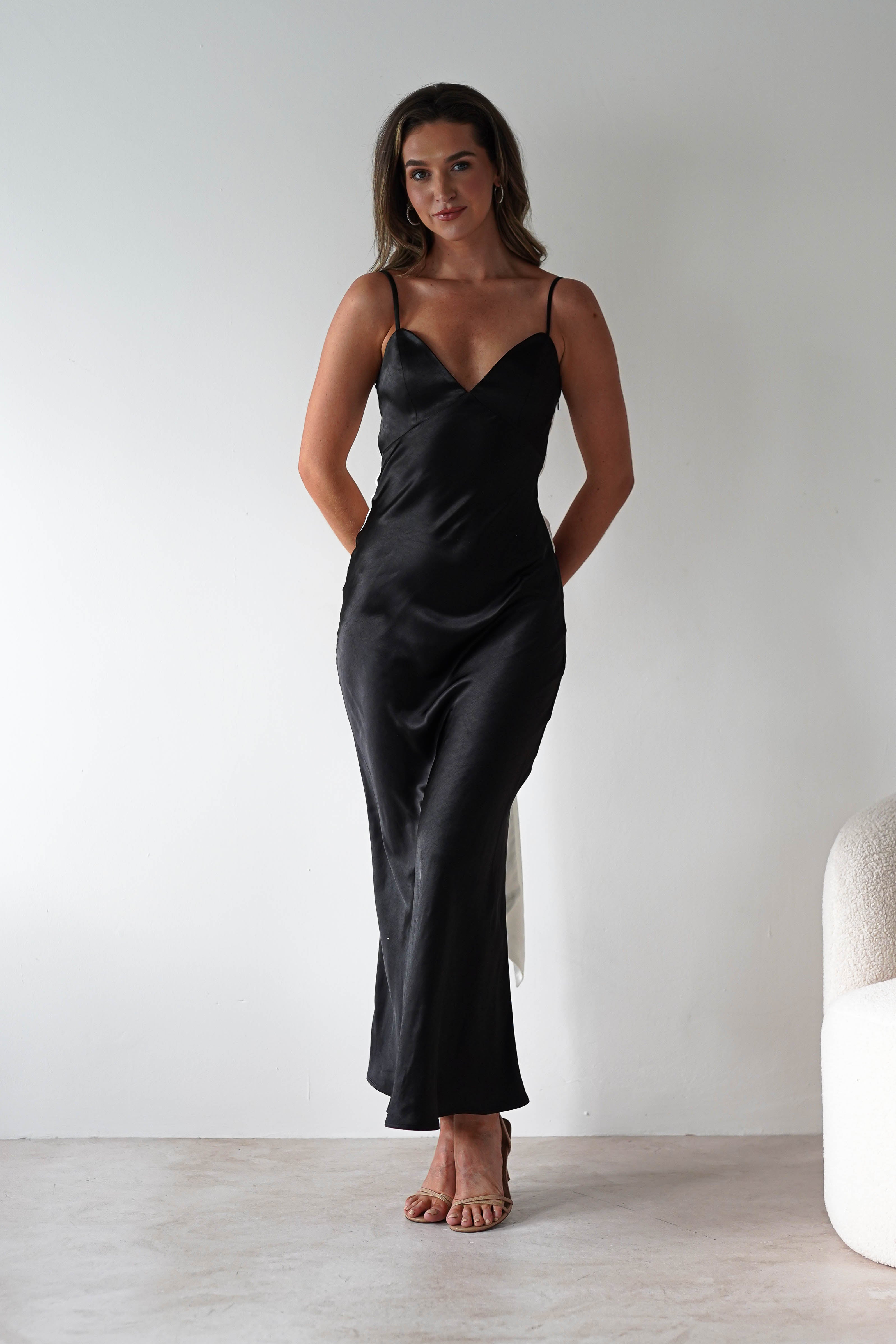 A woman stands against a plain white wall, wearing a satin black midi dress with spaghetti straps and subtle back tie detail—perfect for bridesmaids. She pairs the look with nude heels, long brown hair flowing, hands behind her back. A white textured chair is partially visible.