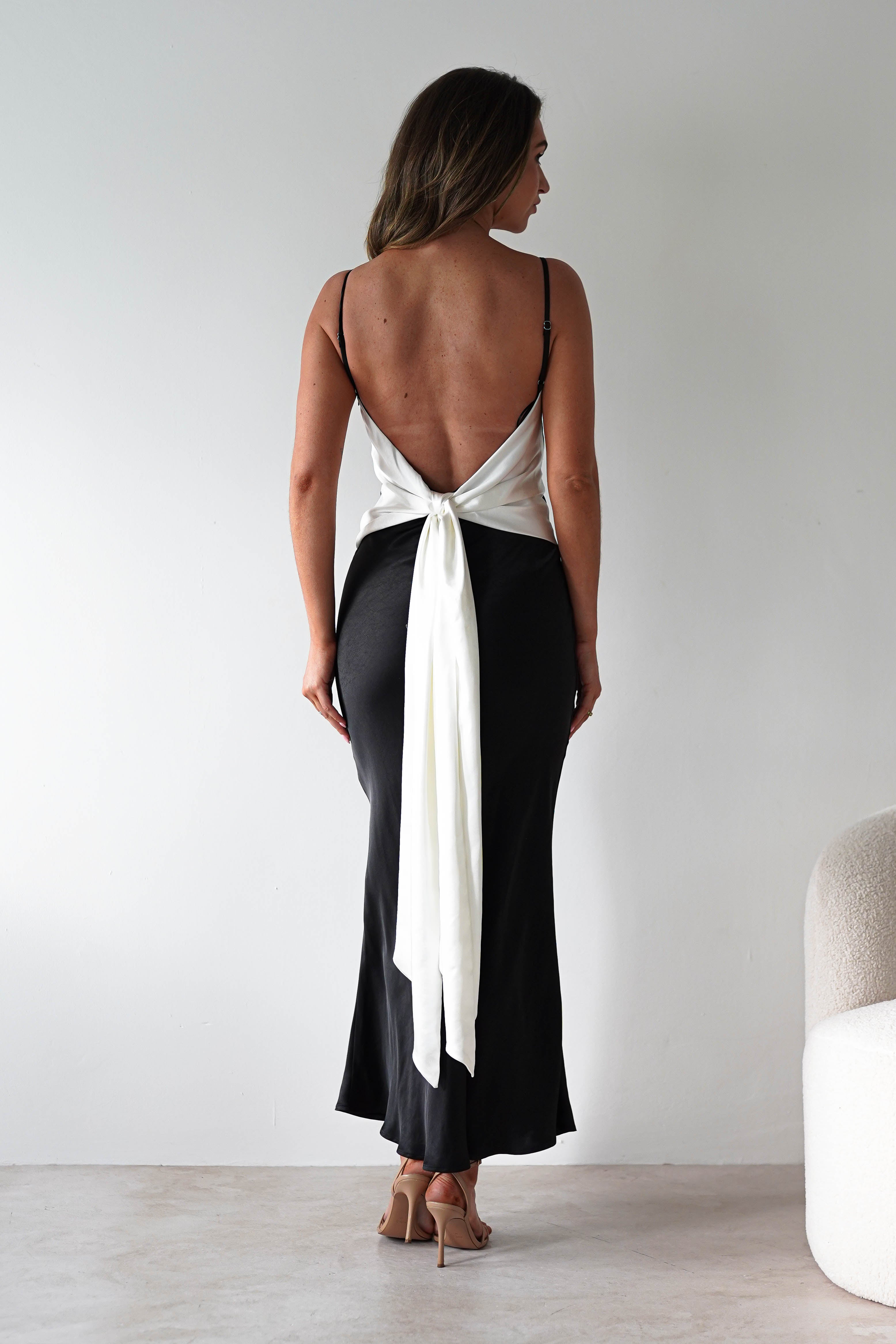 Woman standing indoors facing away, wearing a backless white top with a long back tie detail and a black ankle-length skirt, paired with nude high heels—a chic look perfect for bridesmaids. The minimal setting features light walls and a cream chair.