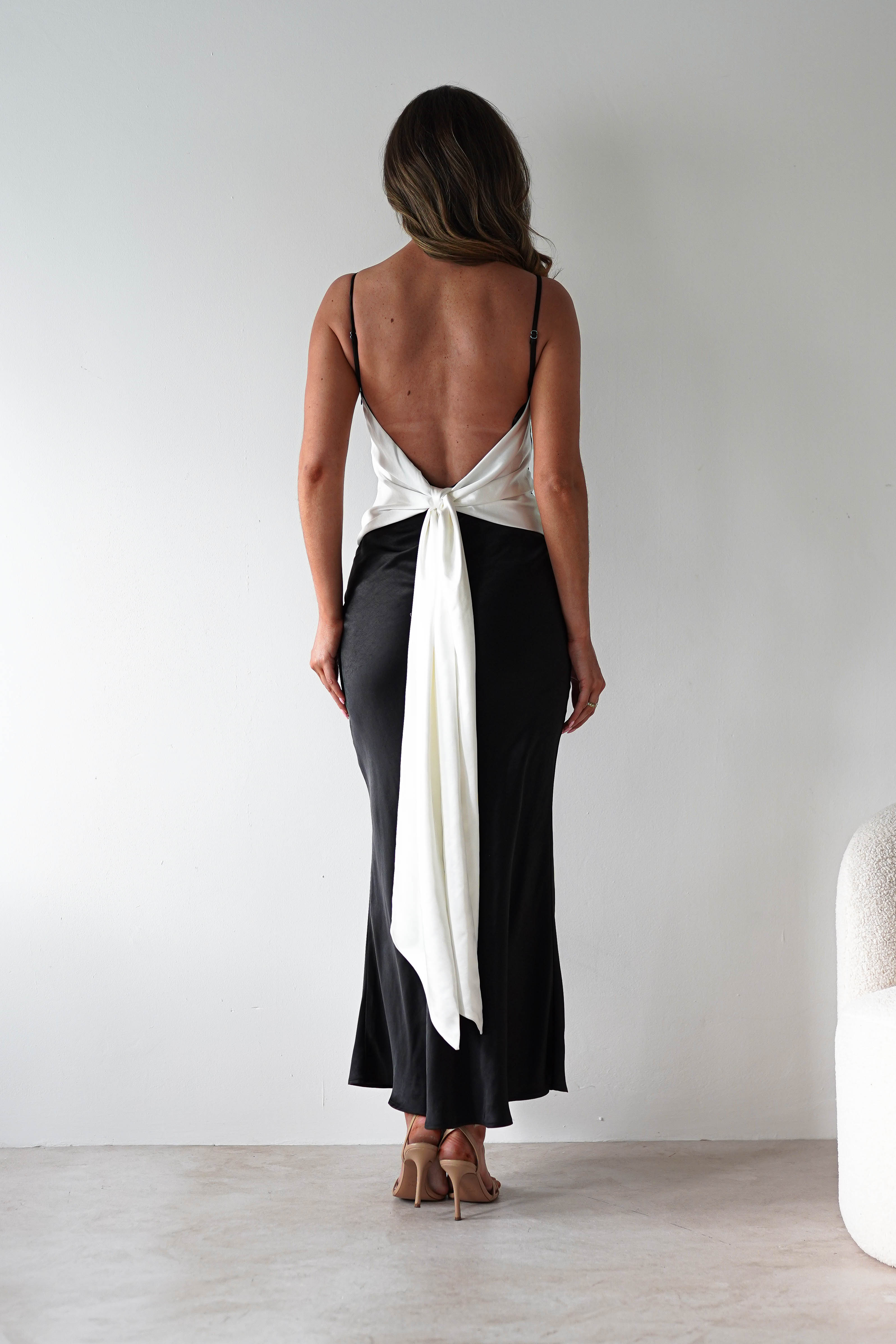 A woman stands facing away in a satin black midi dress with a back tie detail, paired with nude heeled sandals. She’s set against a plain light wall and floor, with part of a textured chair visible on the right—perfect inspiration for bridesmaids style.