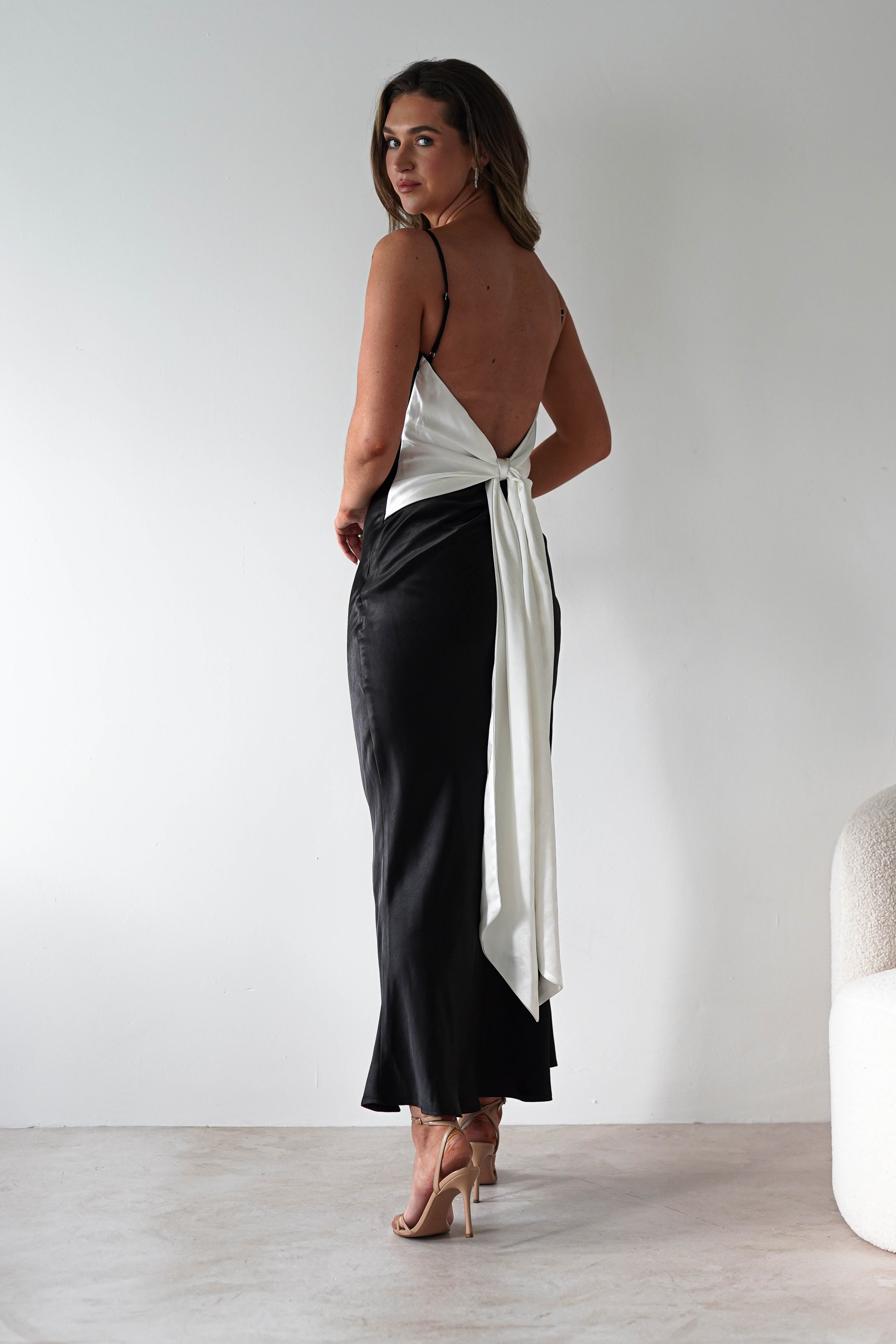 A woman stands indoors in a satin black midi dress with a striking back tie detail and large white bow, paired with nude high-heeled sandals. She looks over her shoulder at the camera, perfect inspiration for stylish bridesmaids.