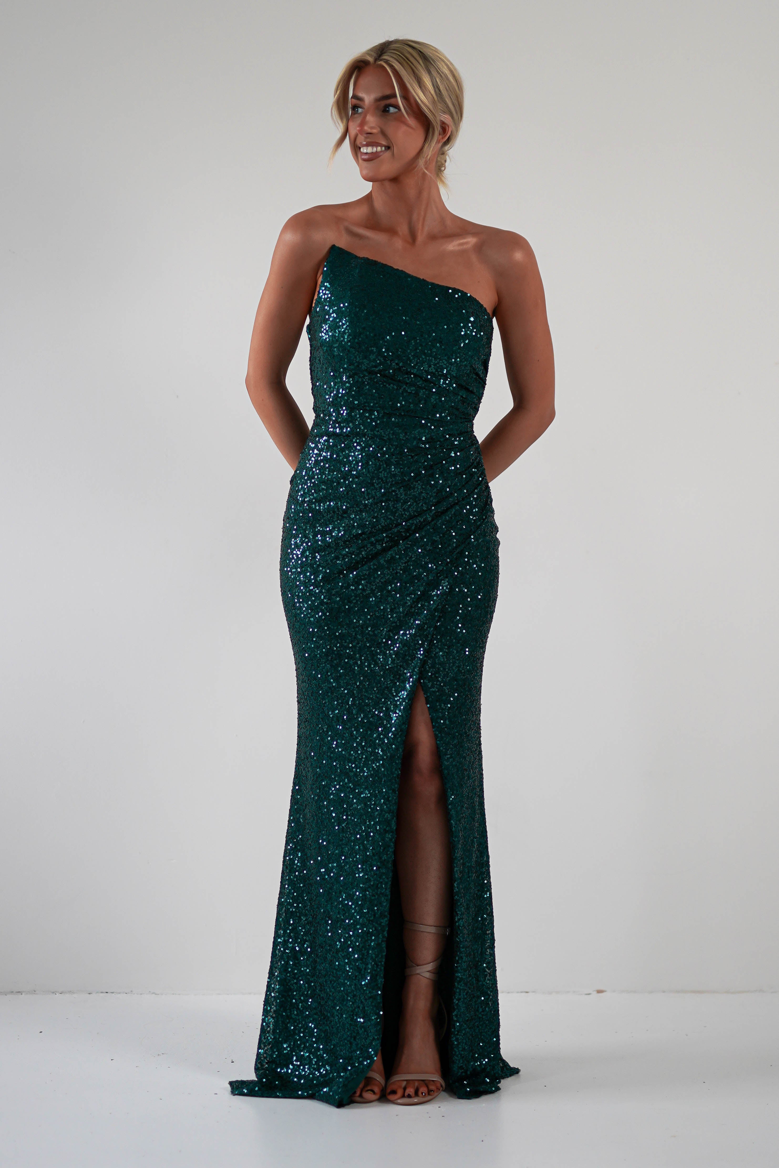 A woman stands against a plain background wearing a strapless sequin gown in dark green with a thigh-high slit, perfect for formal events. She has blonde hair styled up and is smiling with her hands behind her back.