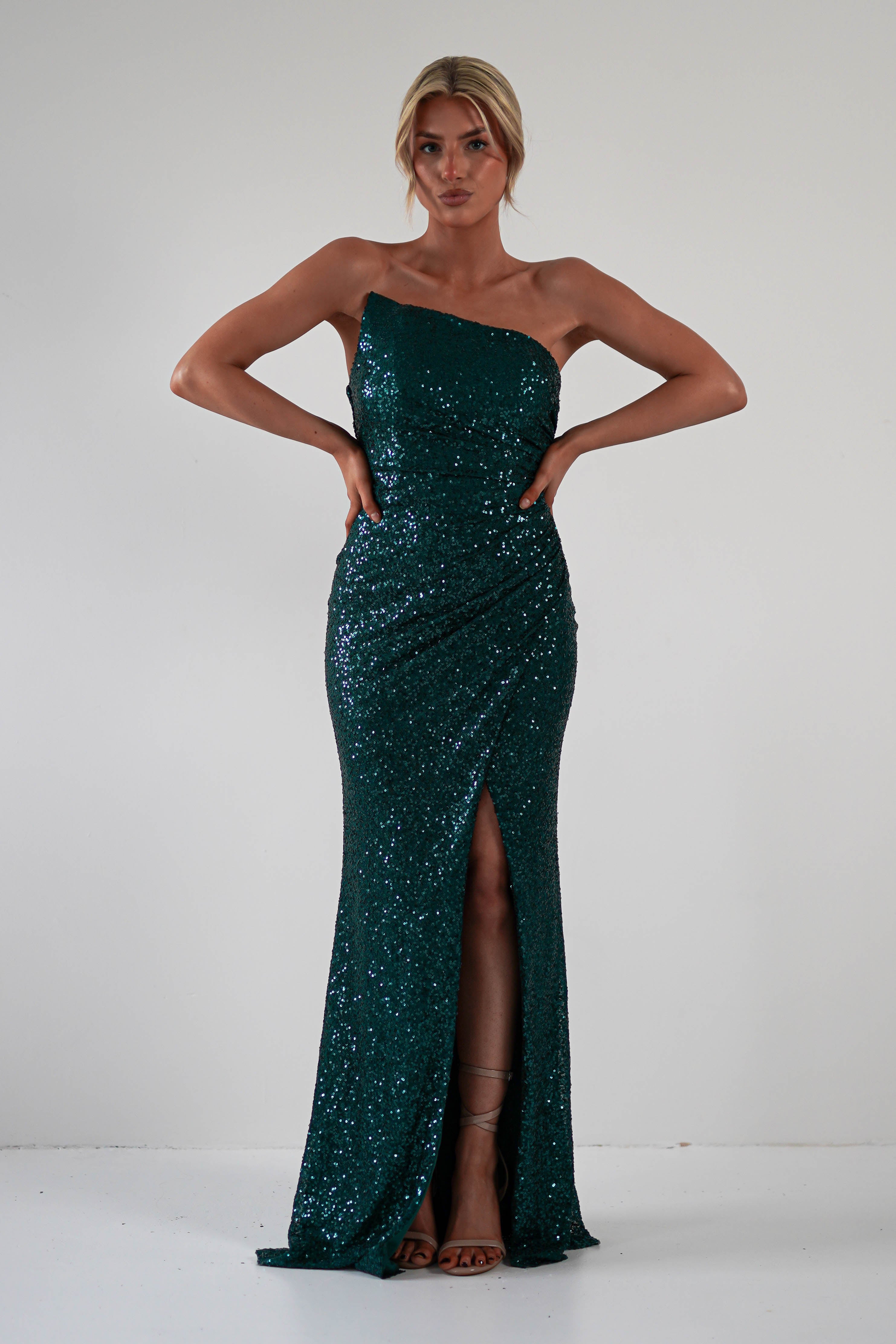 A woman wearing a dark green, one-shoulder sequined gown with a thigh-high slit stands against a plain white background, exuding confidence in attire perfect for formal events. Her hands rest on her hips, showcasing the dresss elegant design.