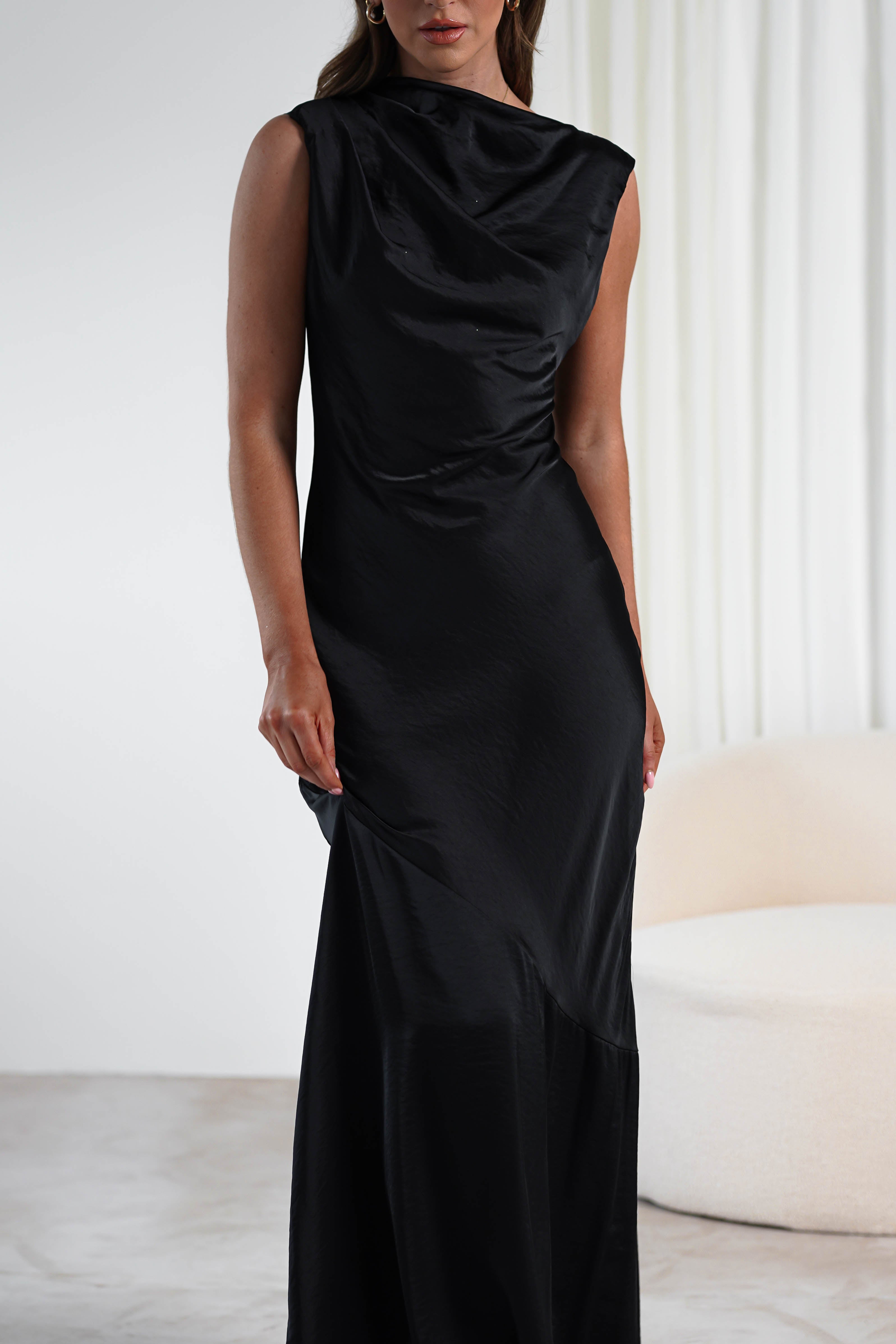 A person wearing a sleek, sleeveless black satin gown stands in a minimalistic room with light-colored walls and a white armchair. Perfect for wedding guests, the dress features a draped neckline and a long, flowing skirt.