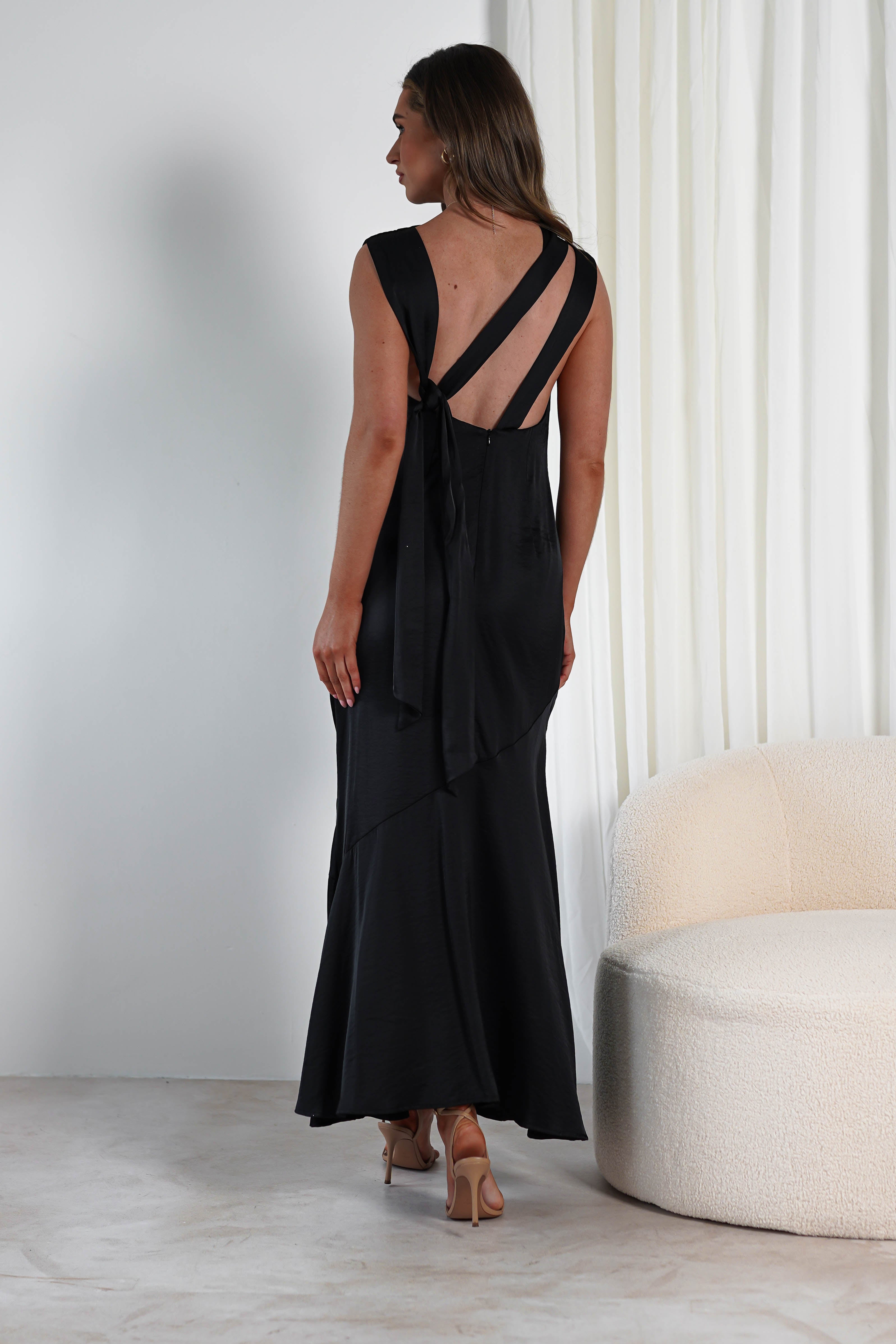 A woman in a black dress with crisscross straps on the back stands beside a white cushioned chair. She gazes to her right in a minimalist room with a white curtain, perfectly poised for an elegant wedding guest appearance.