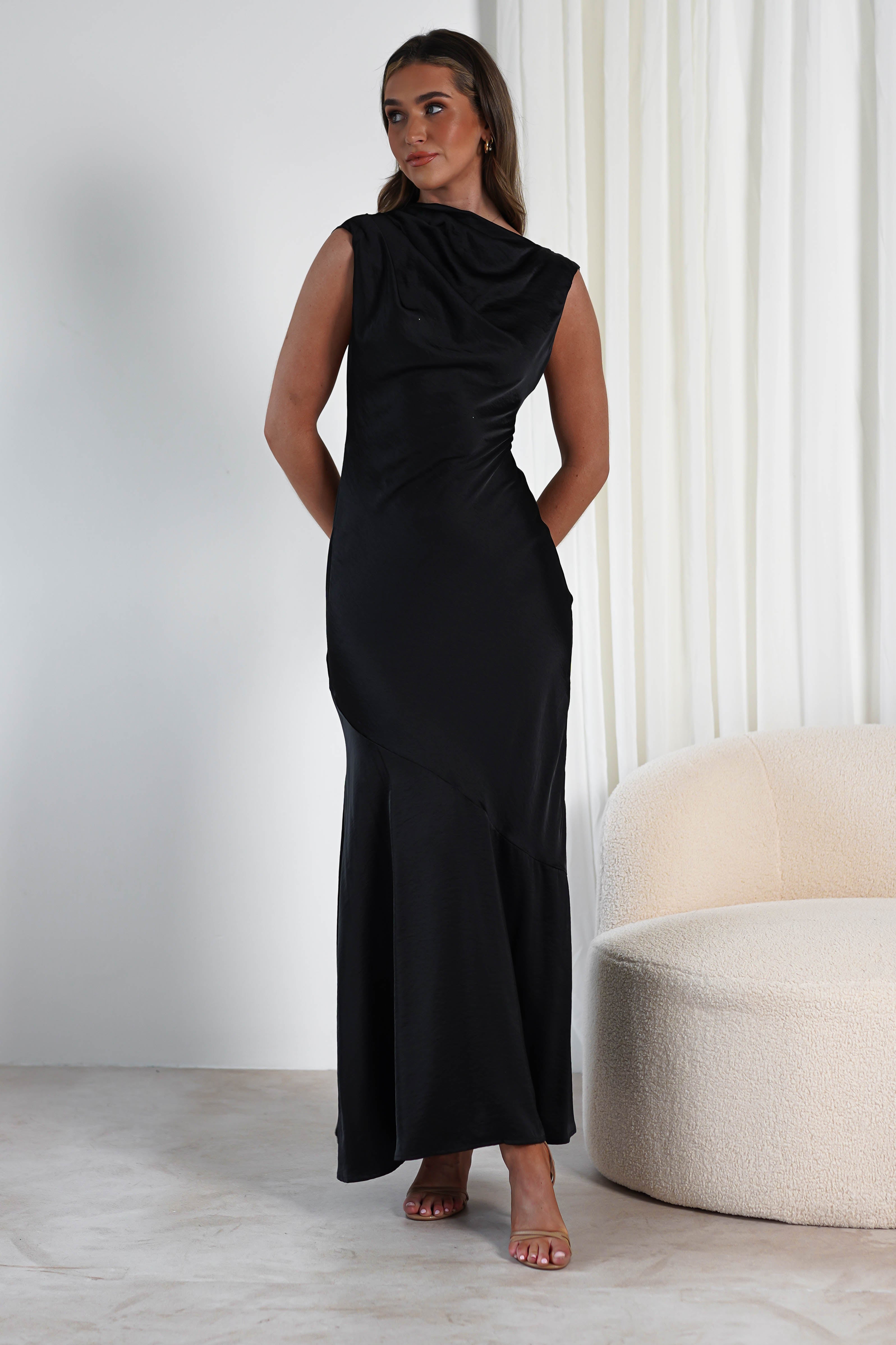 A person wearing a sleek, sleeveless black dress poses elegantly in a modern room, perfect attire for a wedding guest. They stand beside a cream-colored, curved sofa with white drapes in the background. The light-colored floor beautifully complements the minimalist decor.