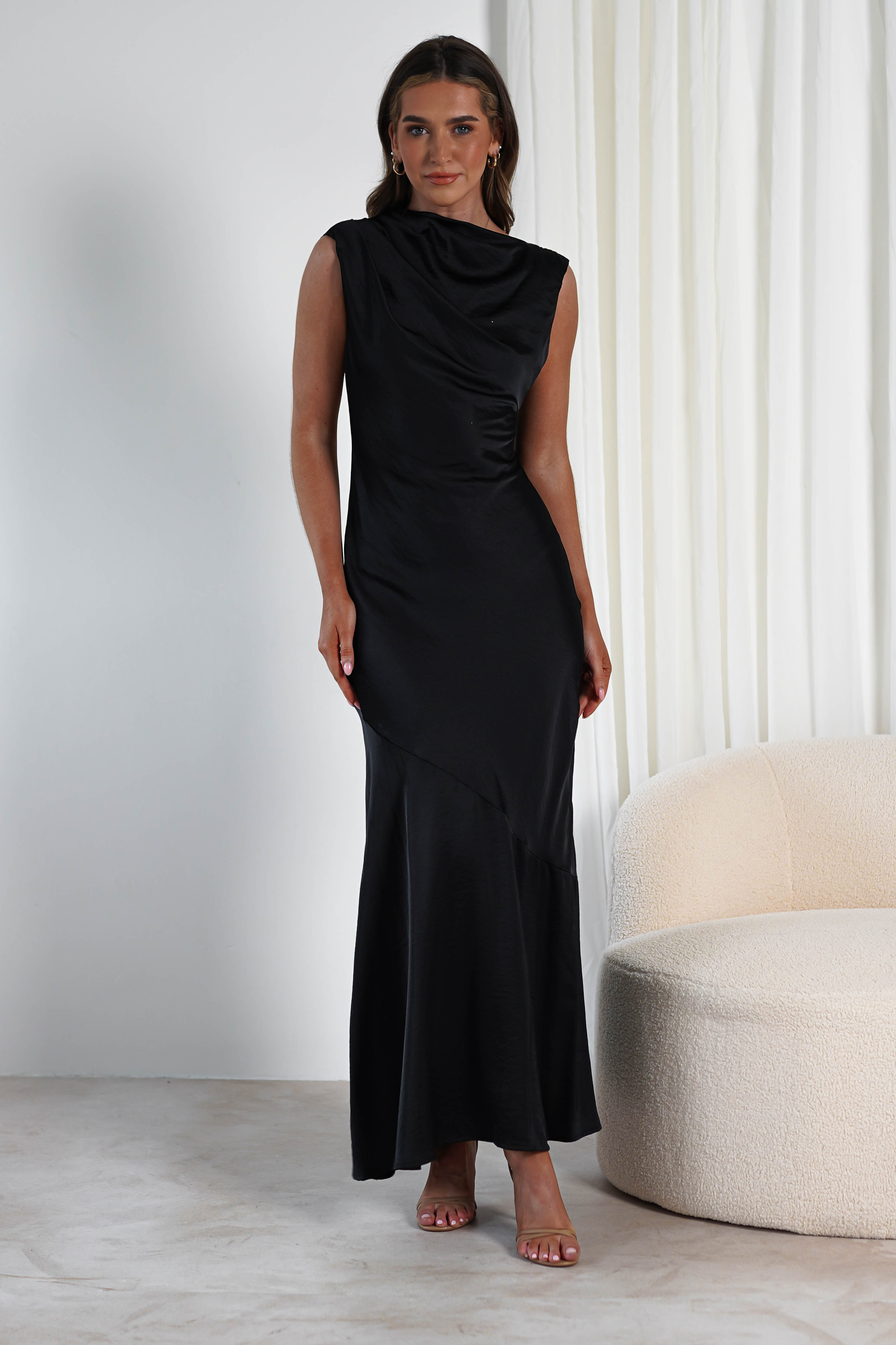 A woman stands confidently in a long, elegant black dress with a draped neckline, perfect attire for a wedding guest. She is indoors next to a light-colored, textured sofa, against a backdrop of white curtains.