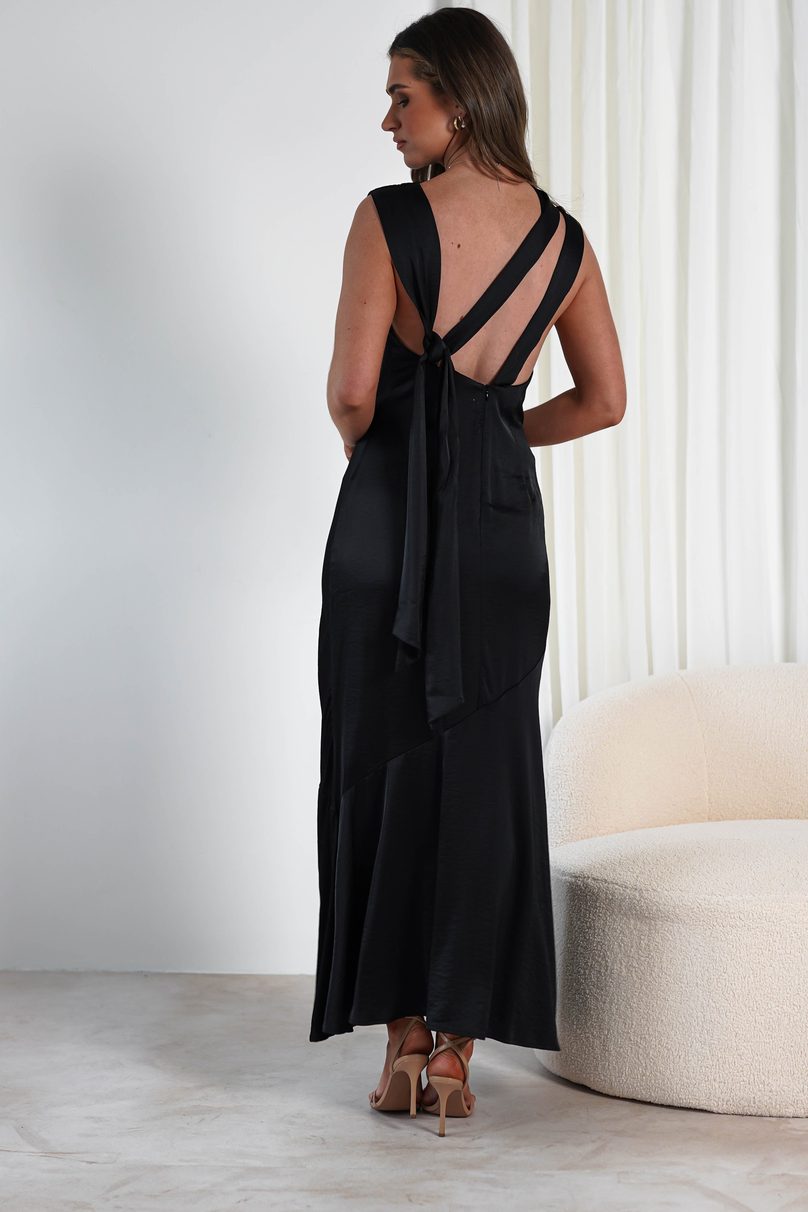 A woman in a black satin dress with crisscross straps and an open back stands facing away, perfect for a wedding guest. The dress features a long bow at the waist. She wears high heels and stands in a room with a cream round chair and white curtains.