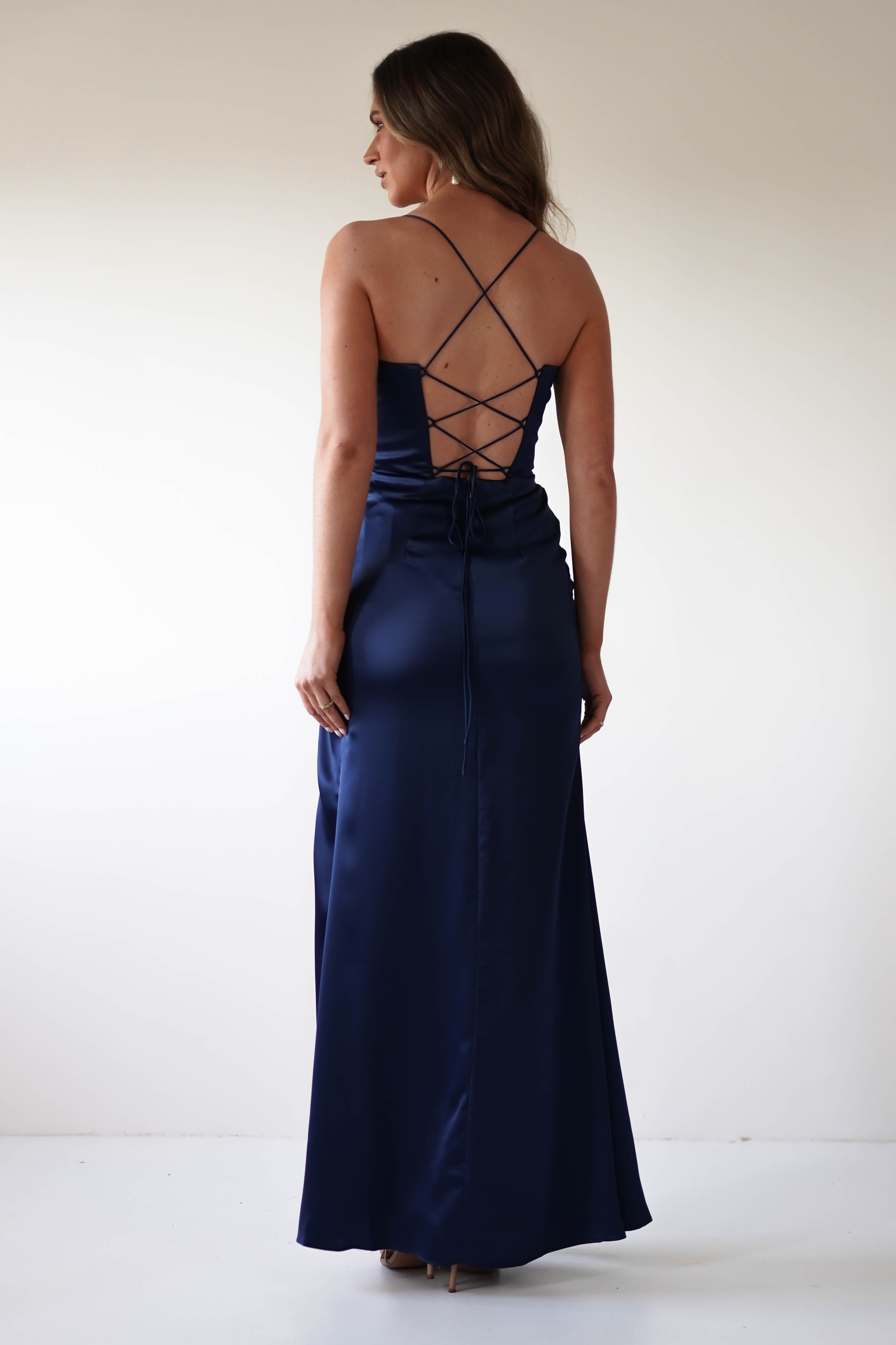 A woman stands wearing a long, navy blue satin corset dress with an open back and crisscross straps. Perfect for any special occasion, she is facing slightly away, her hair cascading down elegantly against a plain white background.