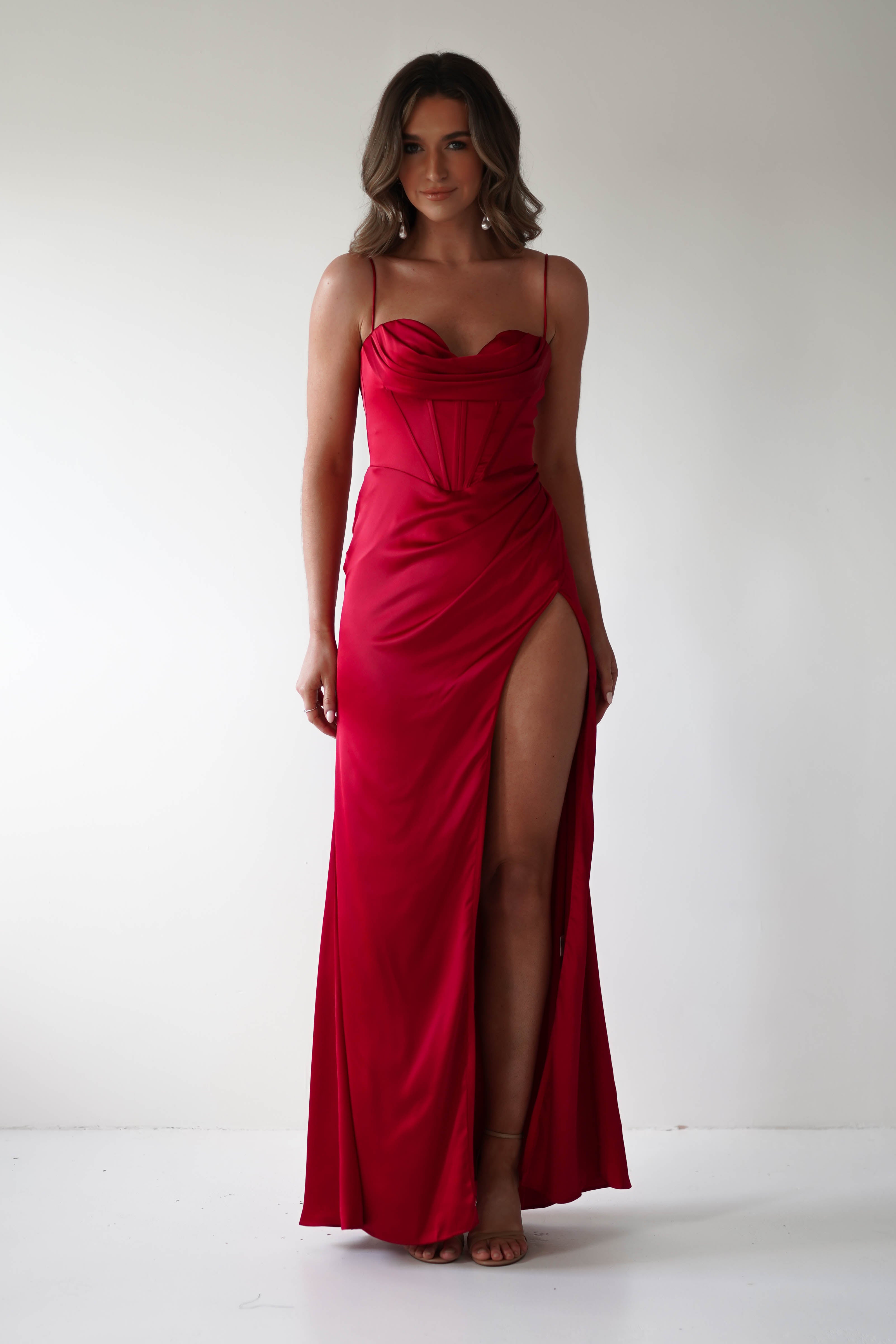 A person wearing a stunning red corset dress with spaghetti straps and a thigh-high slit stands against a plain white background, smiling and posing confidently for this special occasion.