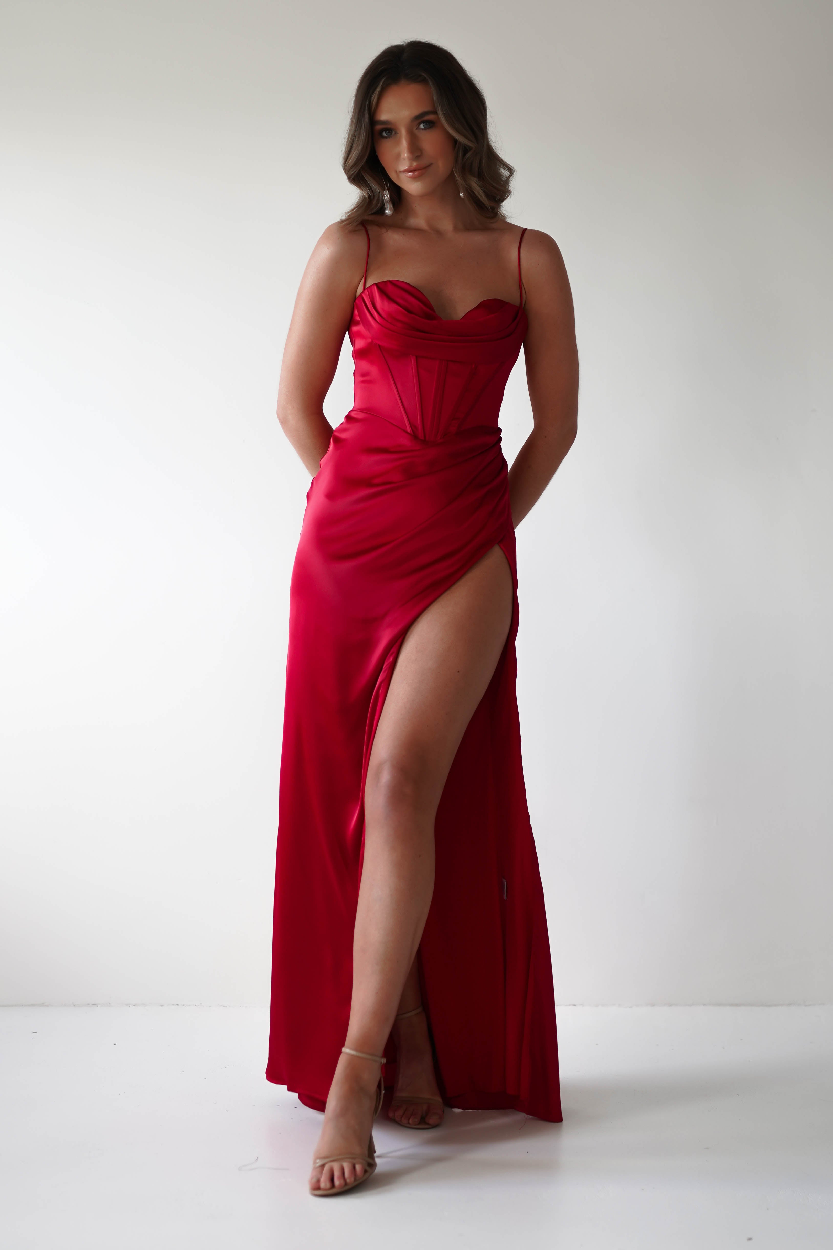 A person stands against a plain white background in a vibrant red, sleeveless corset dress with a thigh-high slit. They have long, wavy hair and are posing with hands behind their back, wearing strappy heels—perfect for any special occasion.