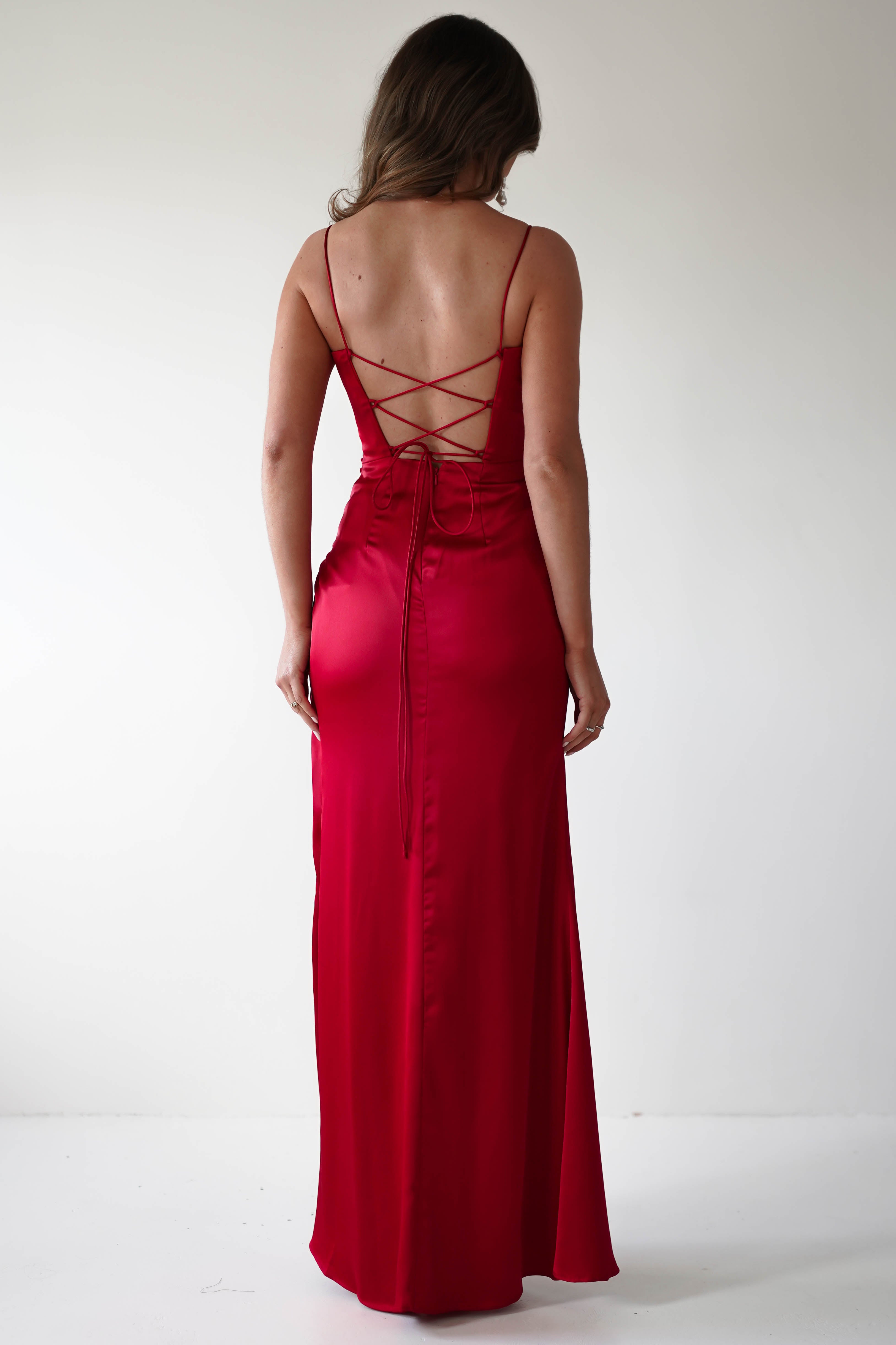 A woman stands against a plain white background, wearing a floor-length, sleeveless red satin gown with a crisscross back, perfect for a special occasion.
